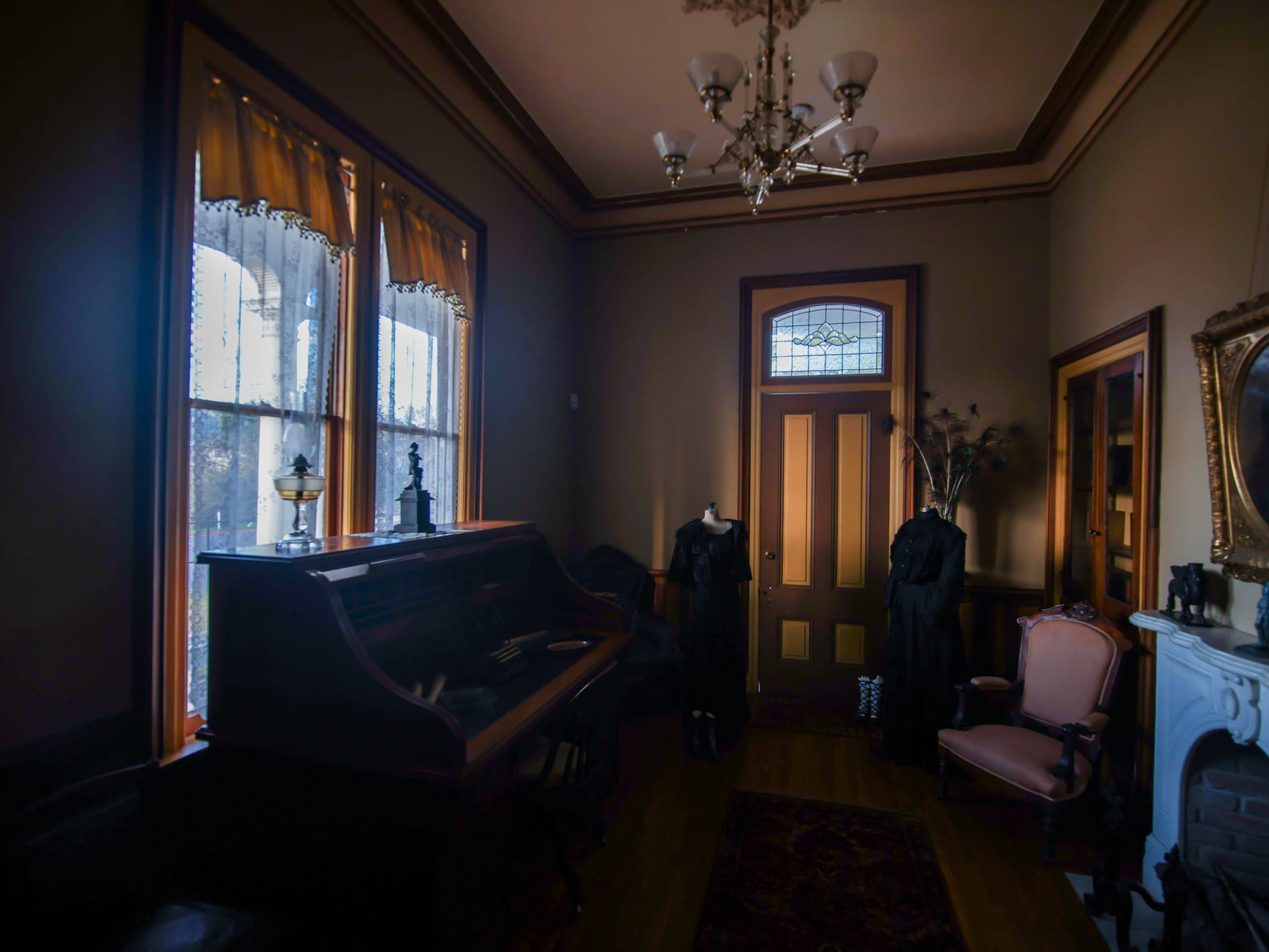 The room features a vintage piano beside a pair of windows with yellow curtains, a wooden door, and a fireplace adorned with a mirror.
