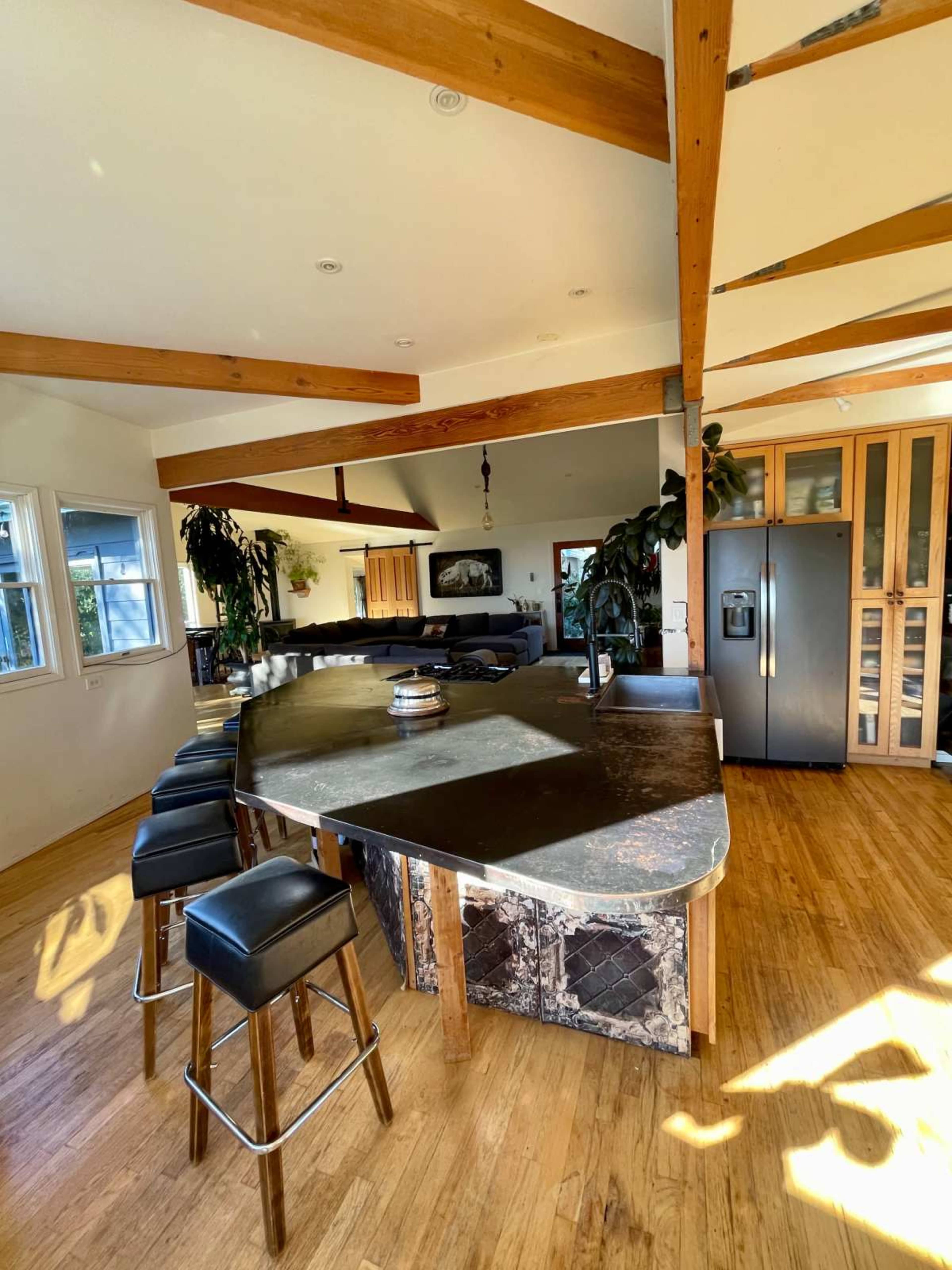 The image shows a modern kitchen with a large curved countertop, wooden beams on the ceiling, and dark cabinetry, featuring a seating area with bar stools.