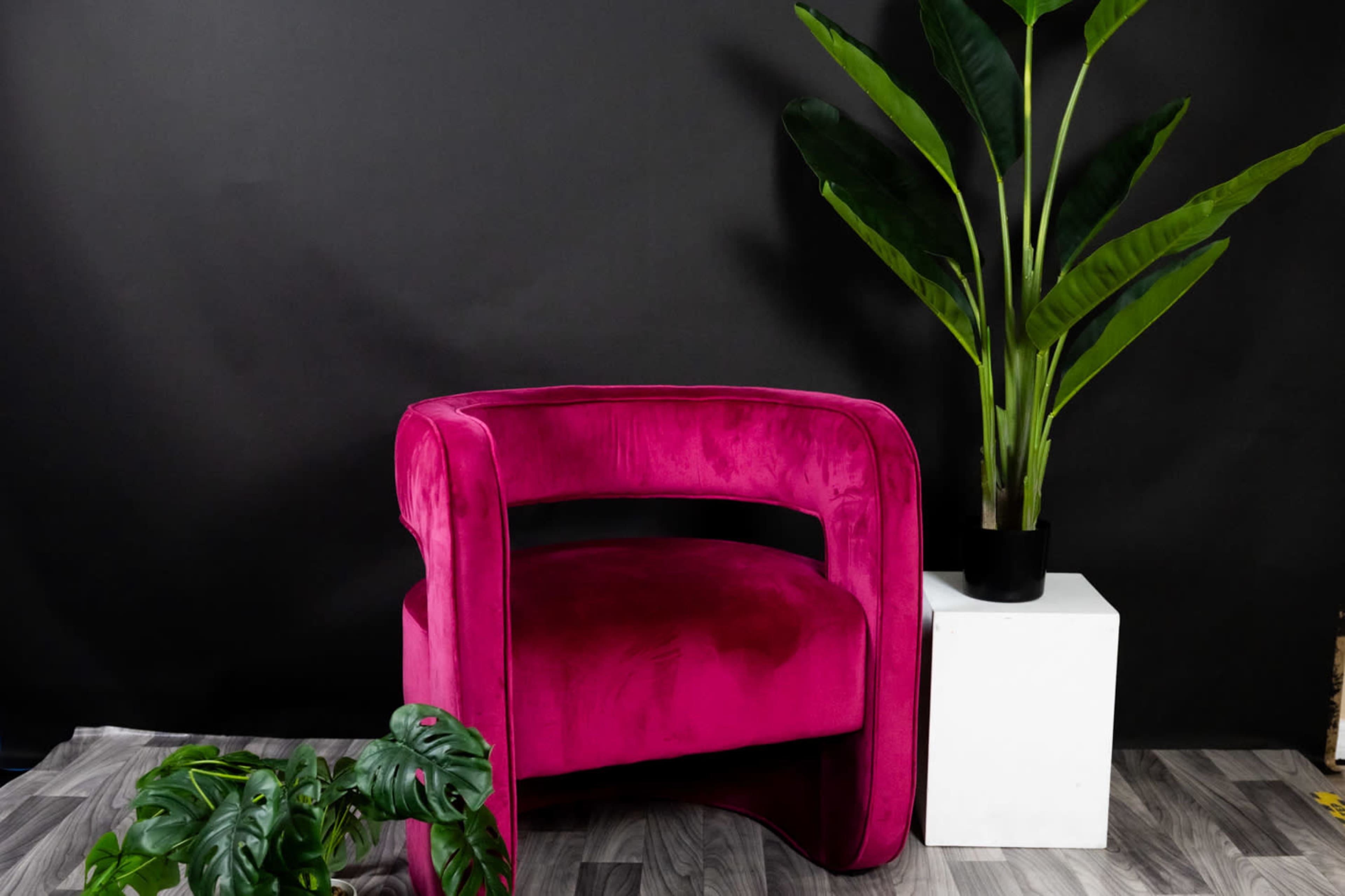 A vibrant pink velvet chair is positioned next to a tall potted plant on a white pedestal against a dark background.