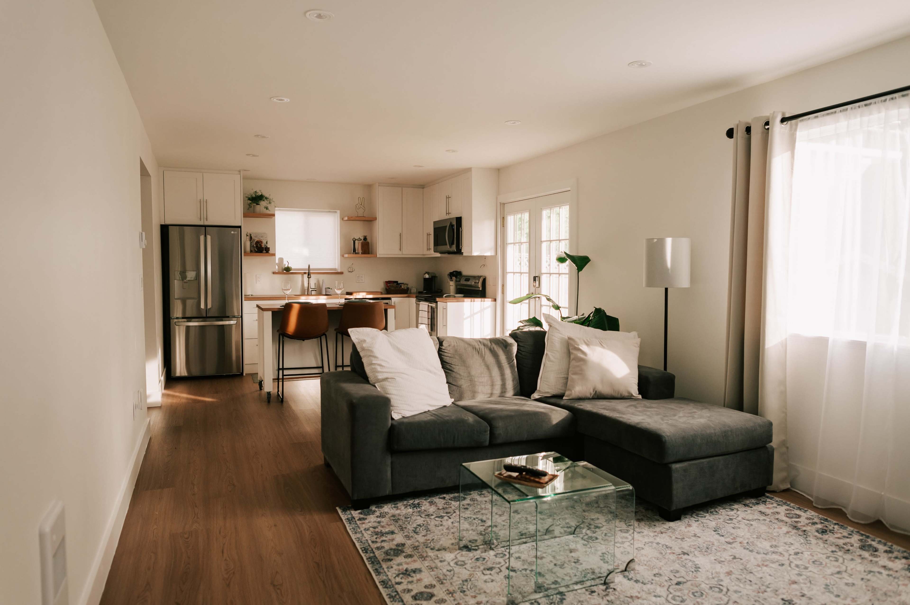 The image shows a modern living room that features a gray sectional sofa, a glass coffee table, and an open kitchen area in the background.