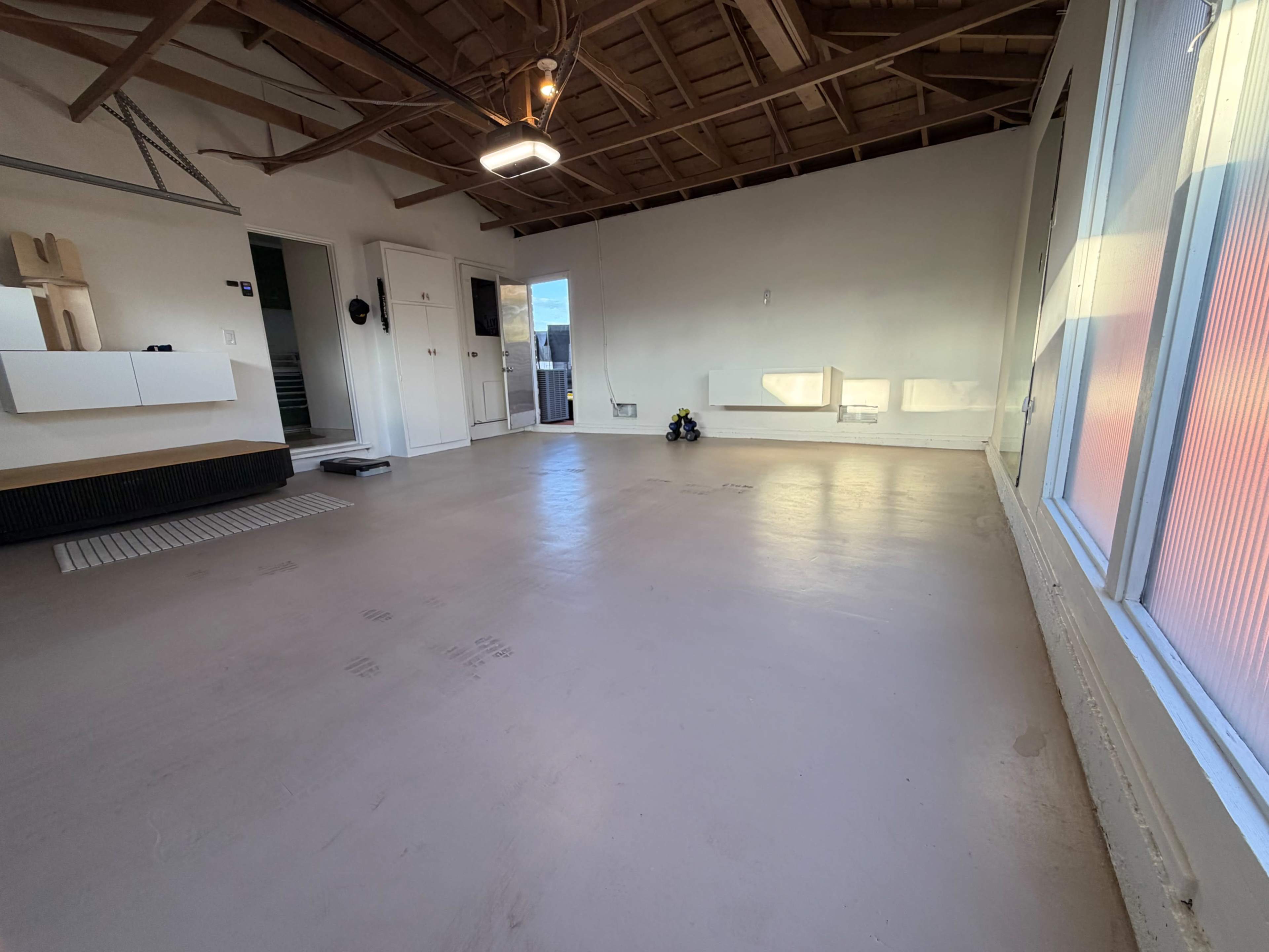The image shows a spacious, empty room with large windows, a concrete floor, exposed wooden beams, and minimal furnishings.