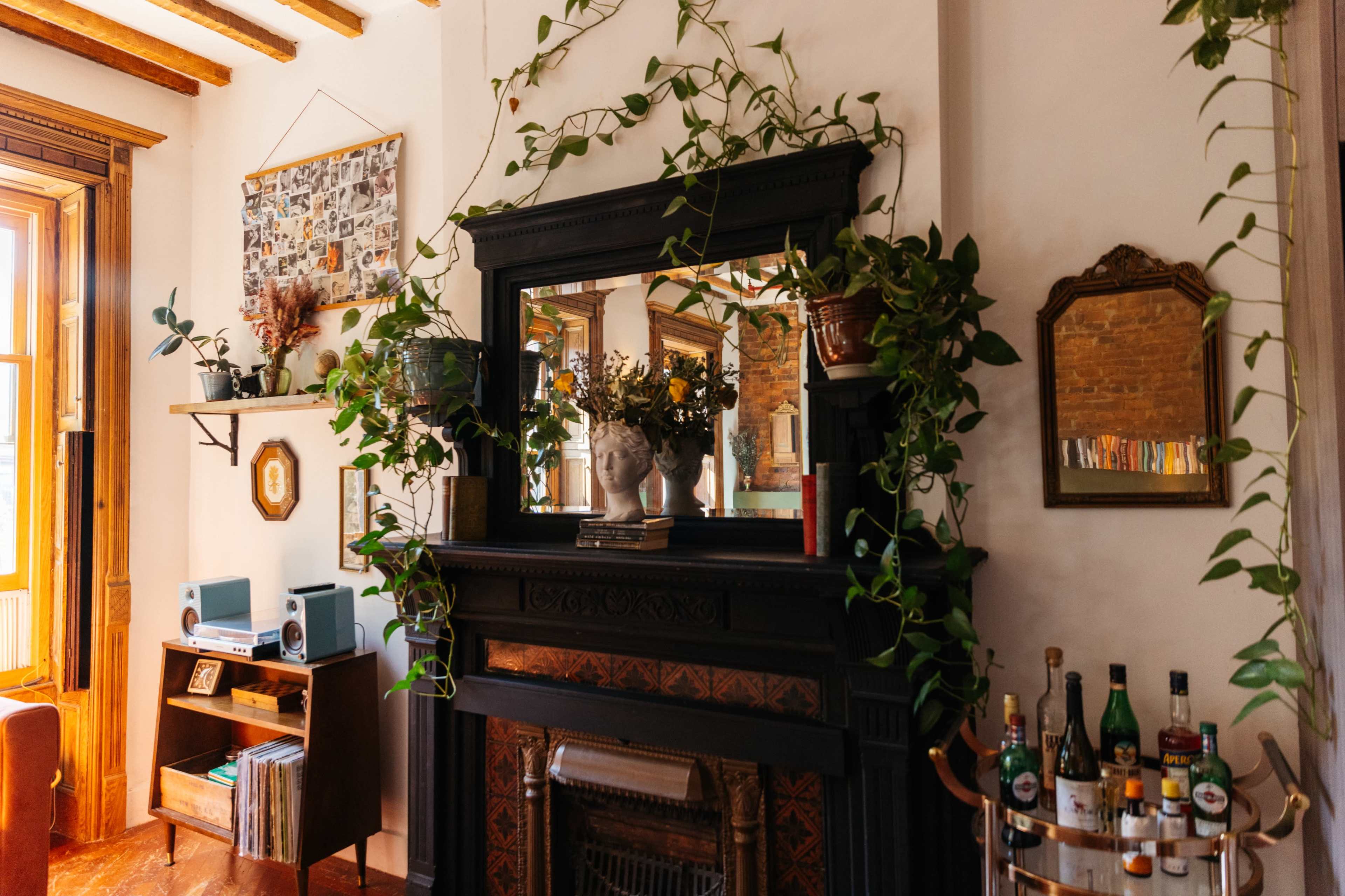 A cozy living room with a black fireplace surrounded by plants, a vintage mirror, framed photos on the wall, and a small bar cart with bottles.