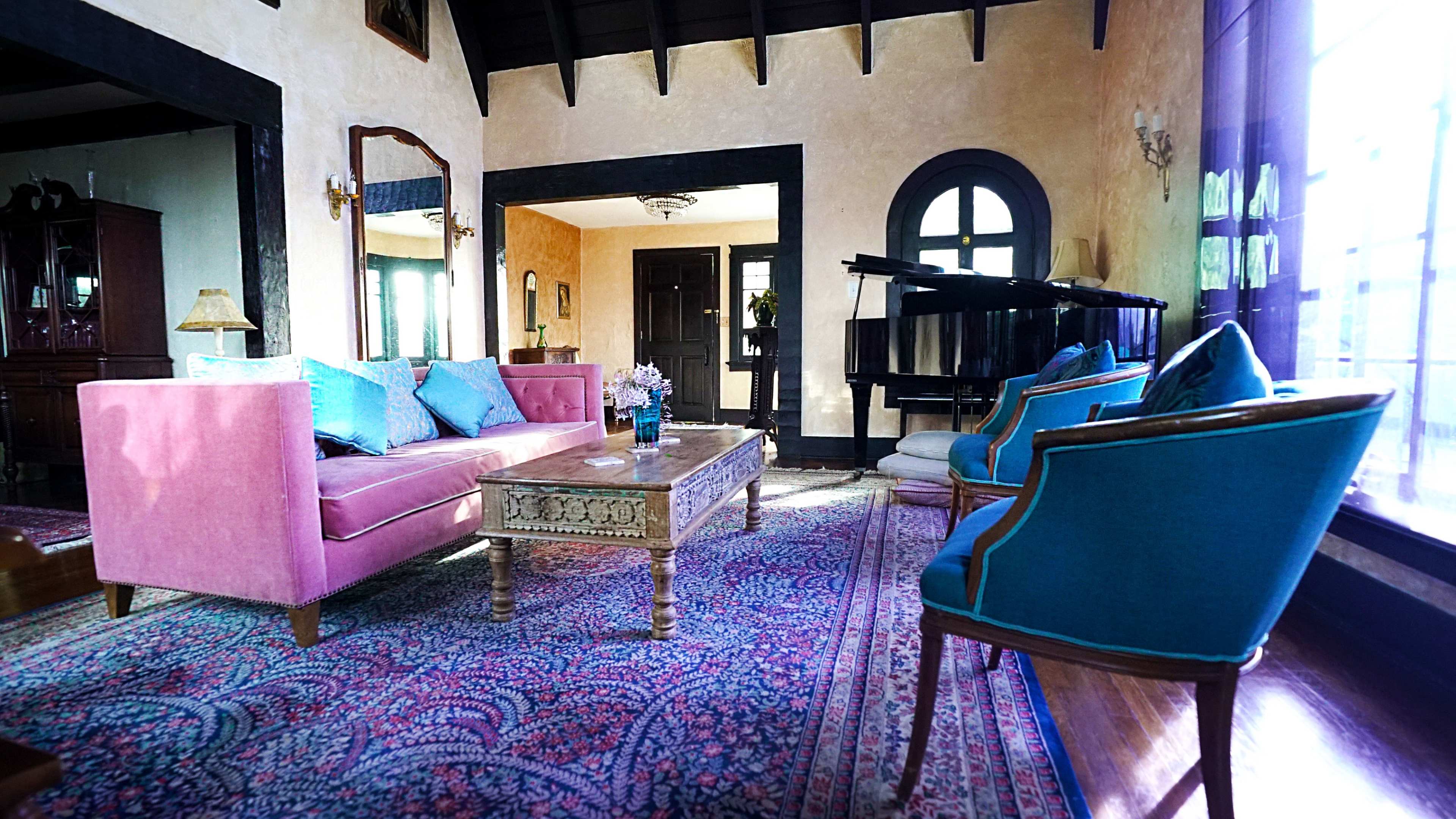 The room features a pink sofa, blue chairs, a decorative coffee table, and a black piano, all set against a backdrop of high ceilings and large windows.