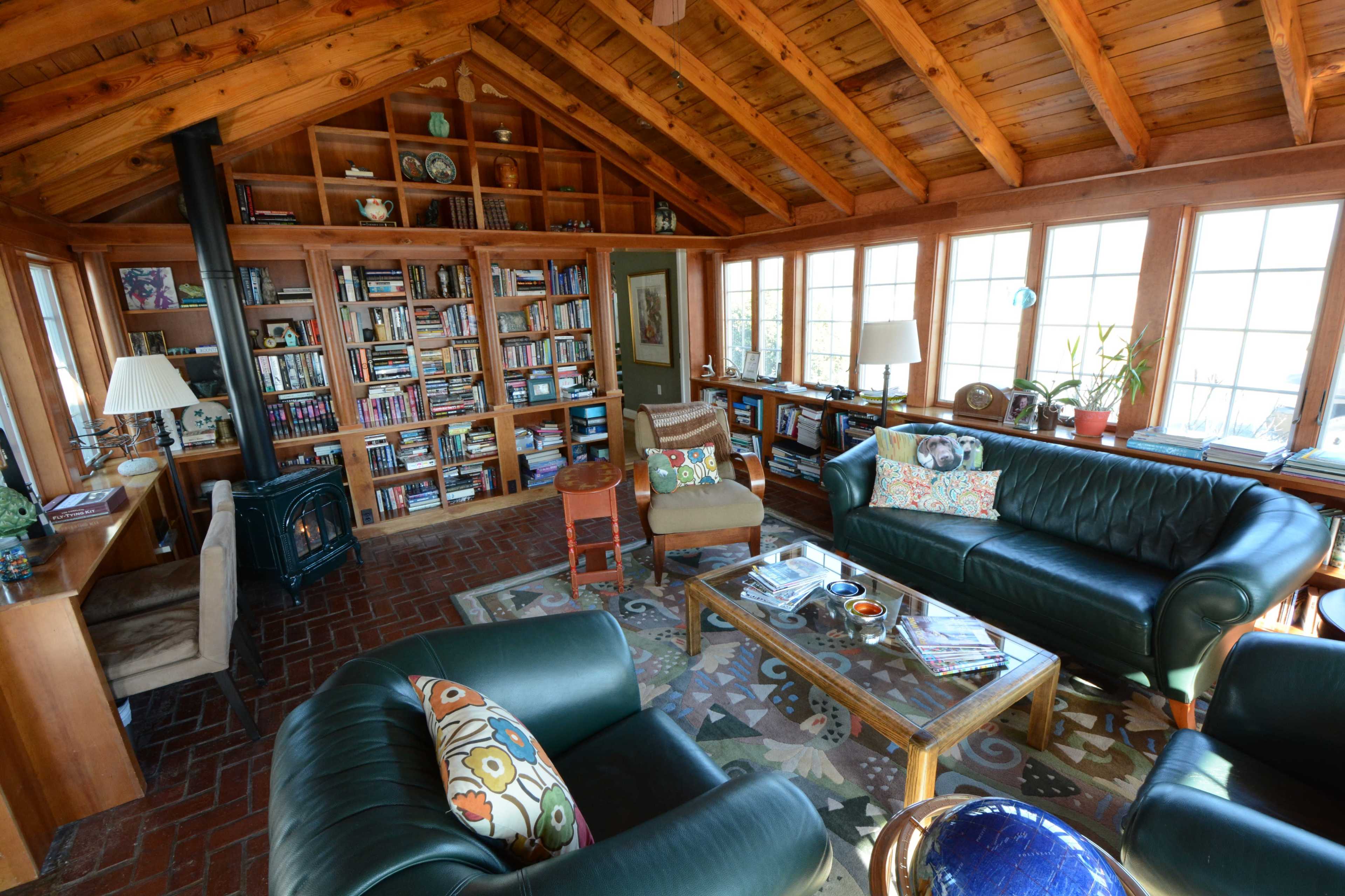 A cozy, wooden sunroom filled with bookshelves, comfortable seating, and large windows providing natural light.