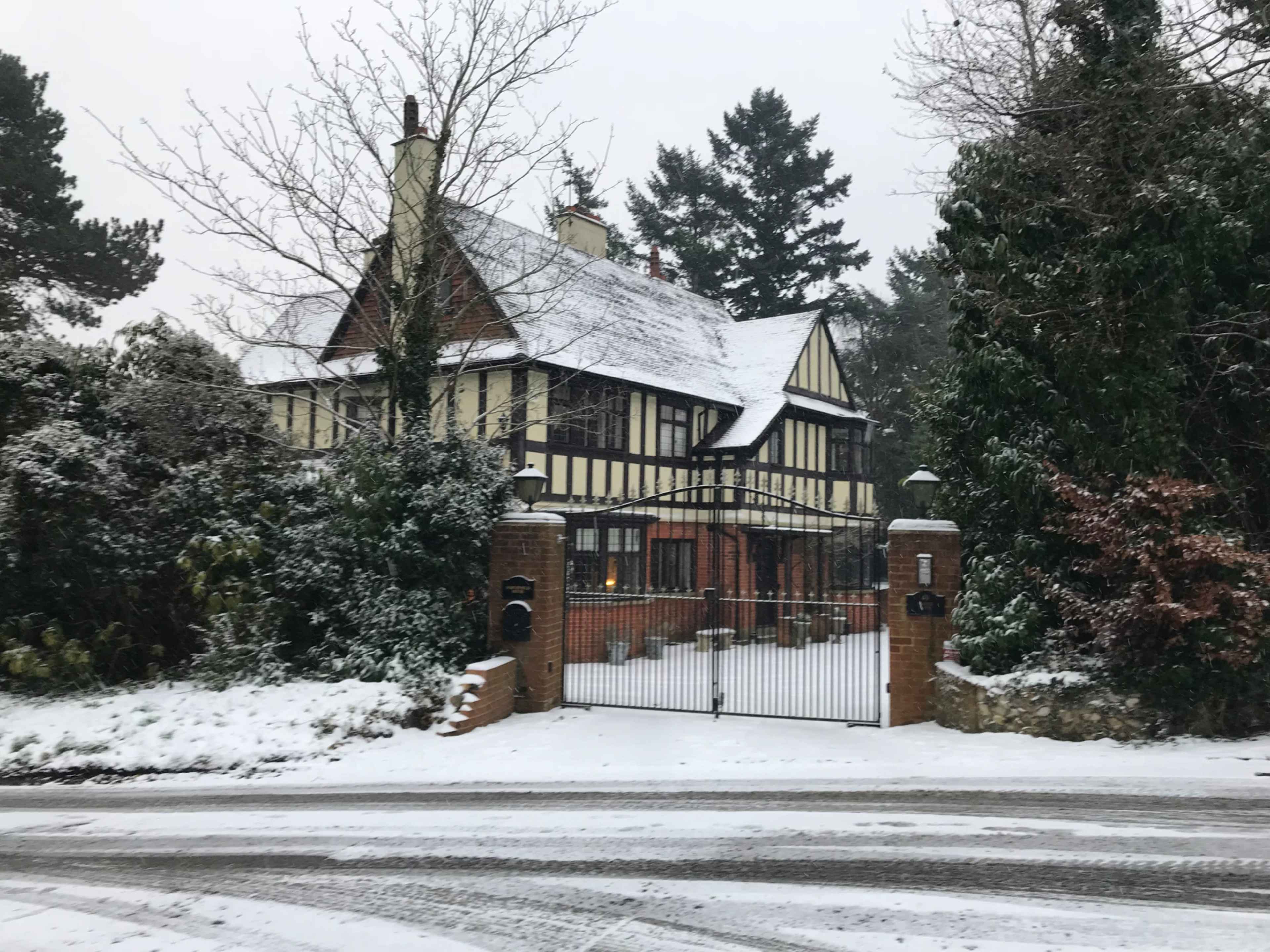 A large, two-story Tudor-style house with a snow-covered yard and a gated entrance is visible in a winter scene.