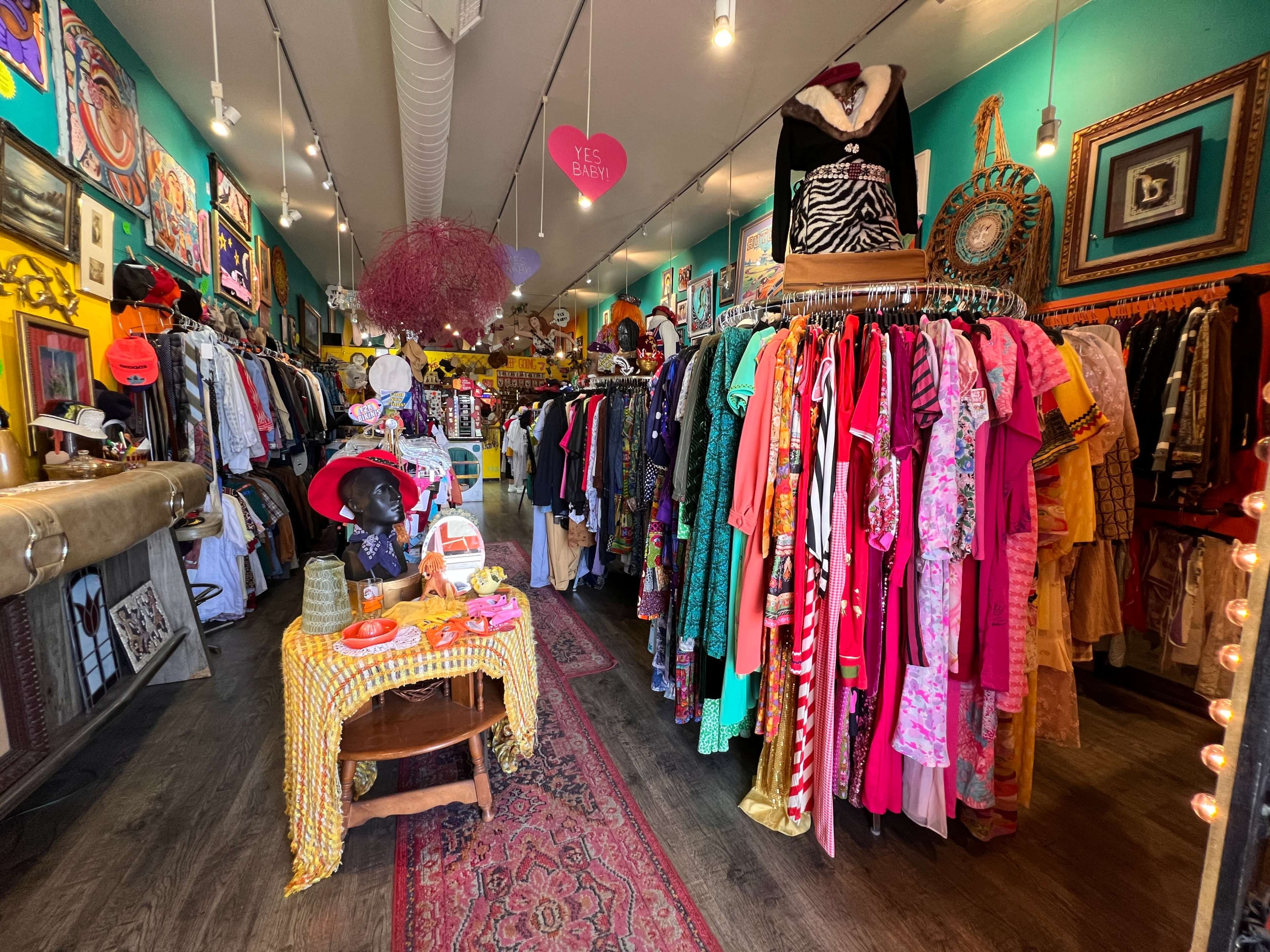 The image shows a colorful boutique filled with a variety of clothing racks, vintage decor, and a small seating area with a table at the front.