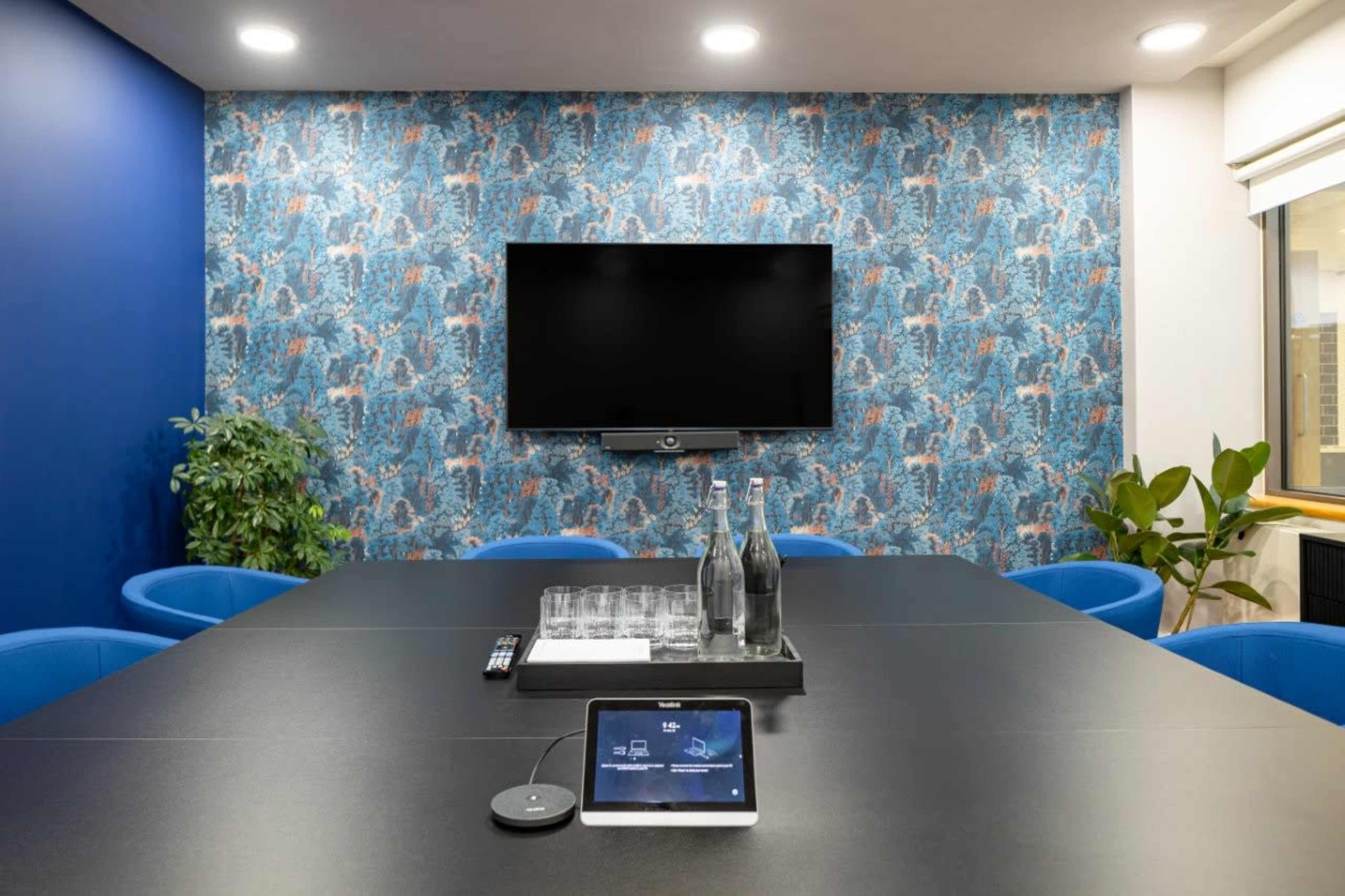 A modern conference room features a large table, blue seating, a television on the wall, and a tablet on the table for presentations.