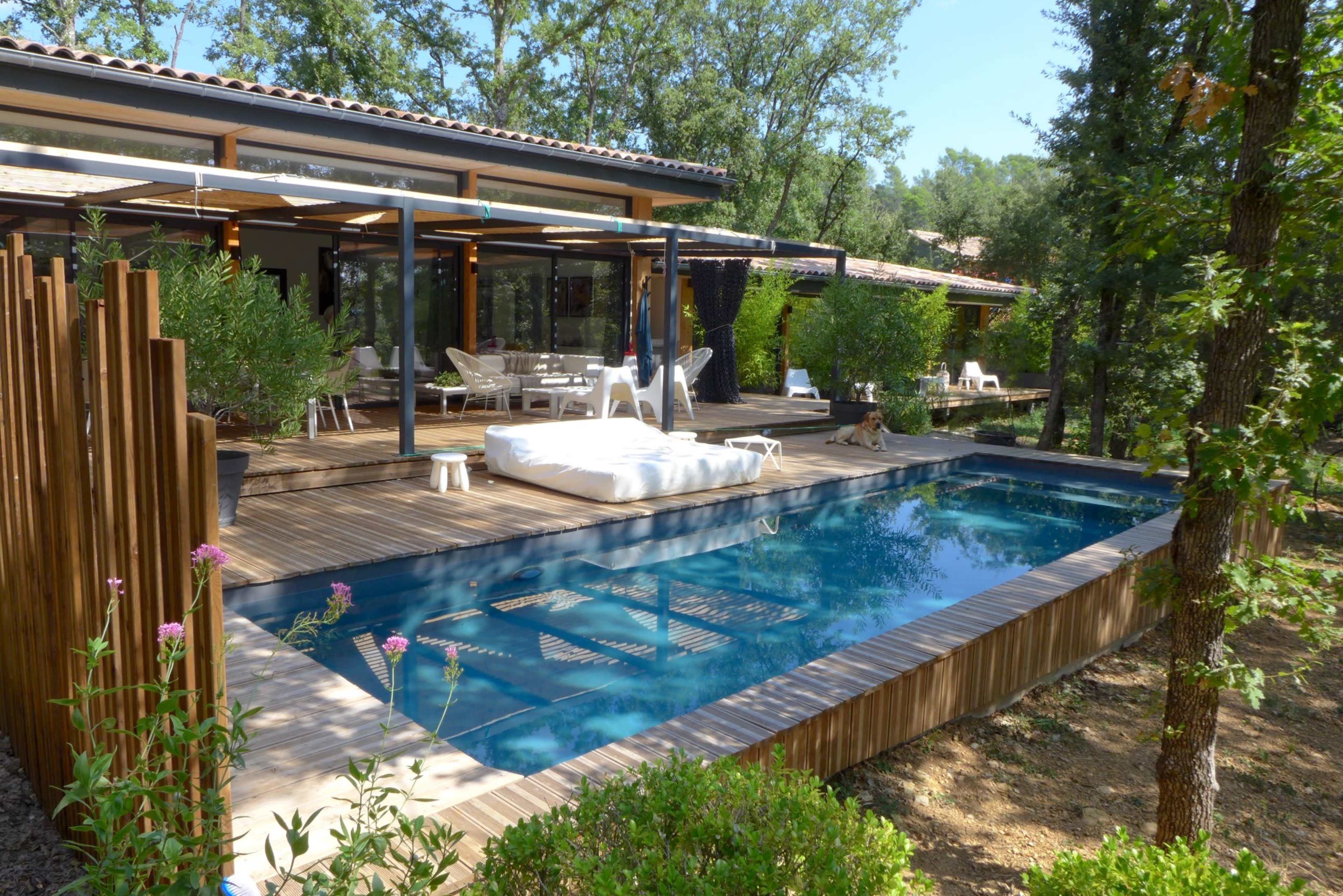 A modern outdoor area featuring a wooden deck, a swimming pool, and a lounge space surrounded by greenery.