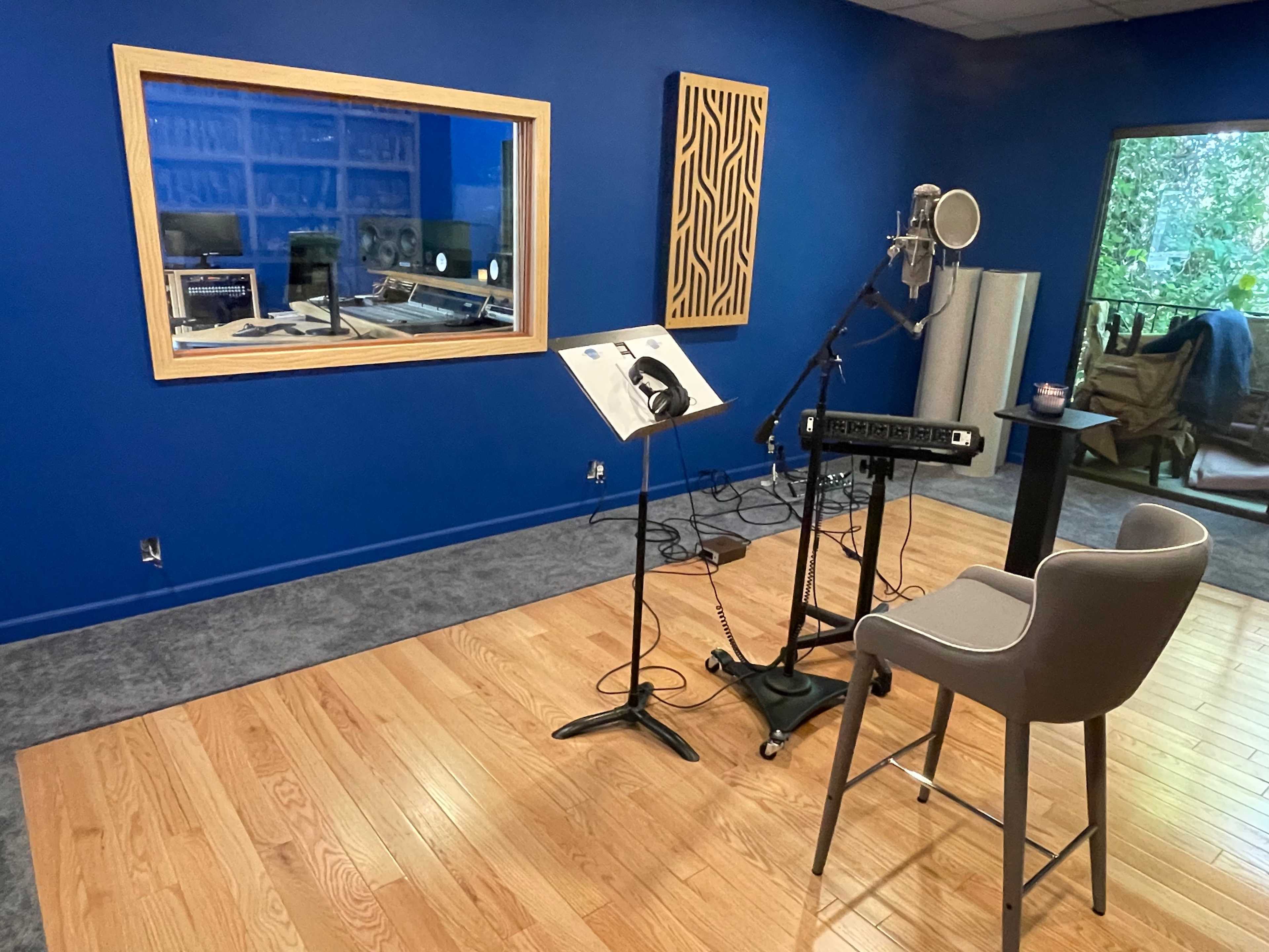 A sound recording studio with a blue wall, a microphone stand, a music stand, and a chair in front of a soundproof glass window.