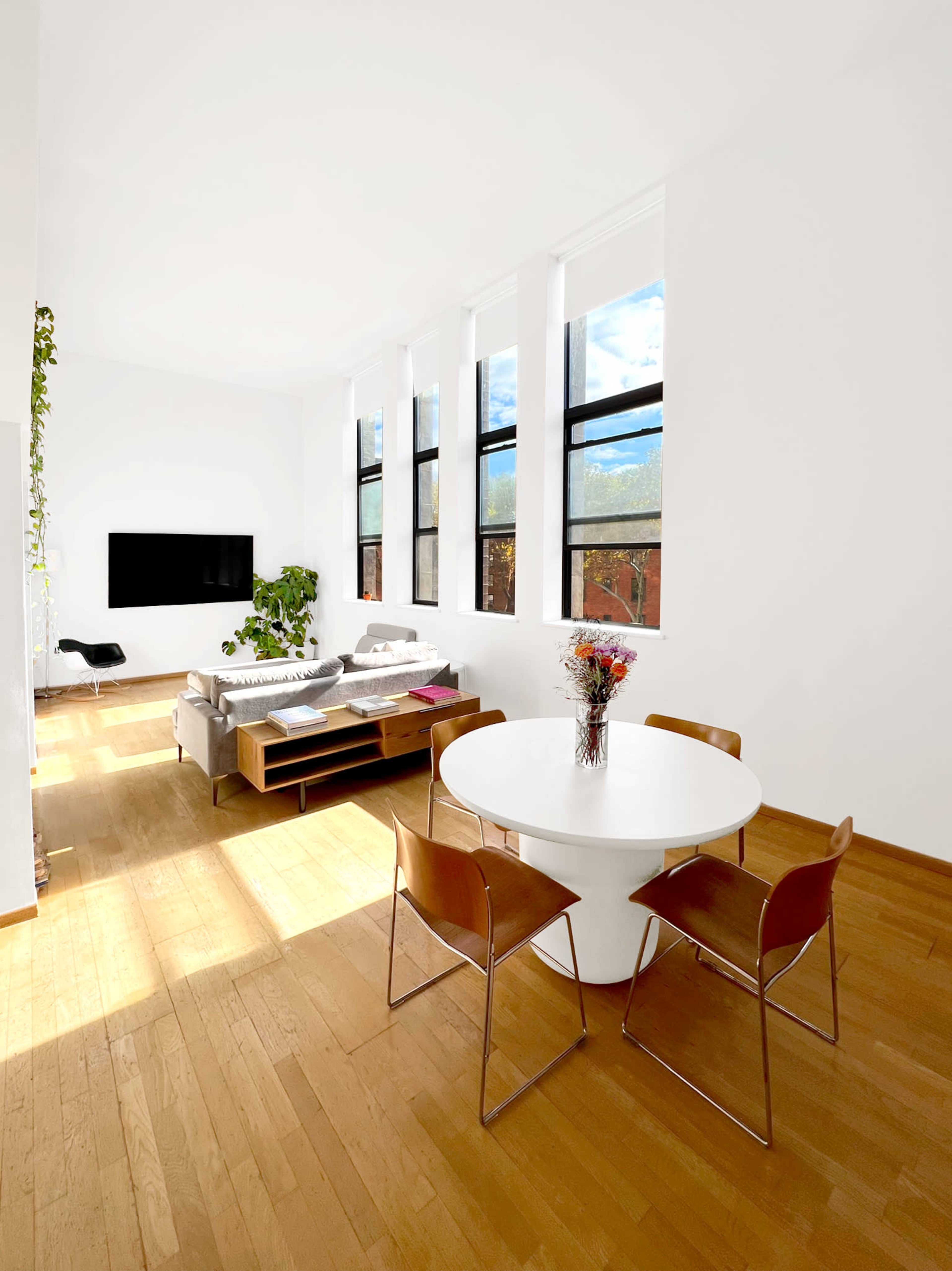 The image shows a modern living space with a round dining table and four chairs, a sofa, a television, and large windows letting in natural light.