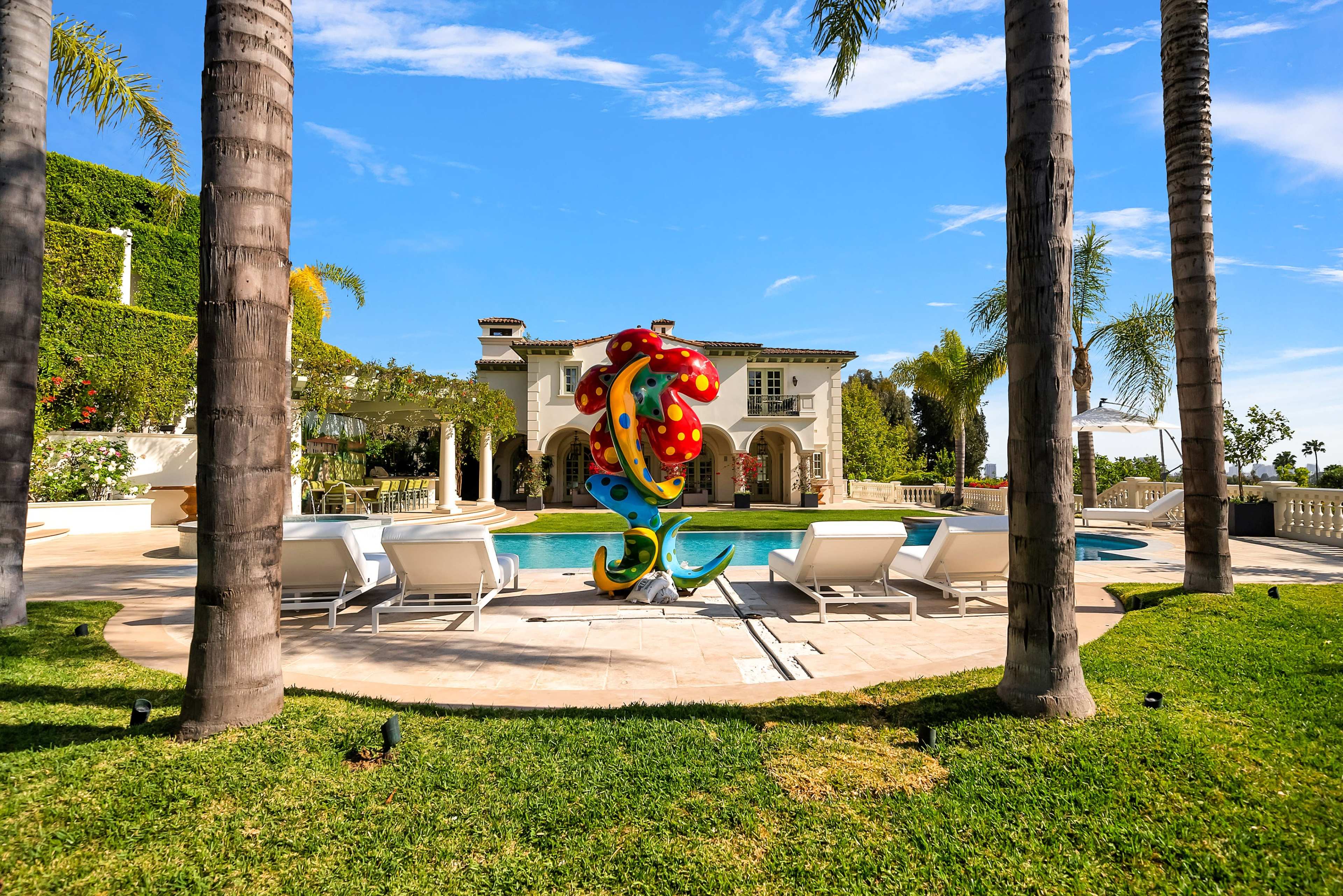 A colorful sculpture stands near a swimming pool surrounded by lounge chairs and palm trees in a luxurious backyard.
