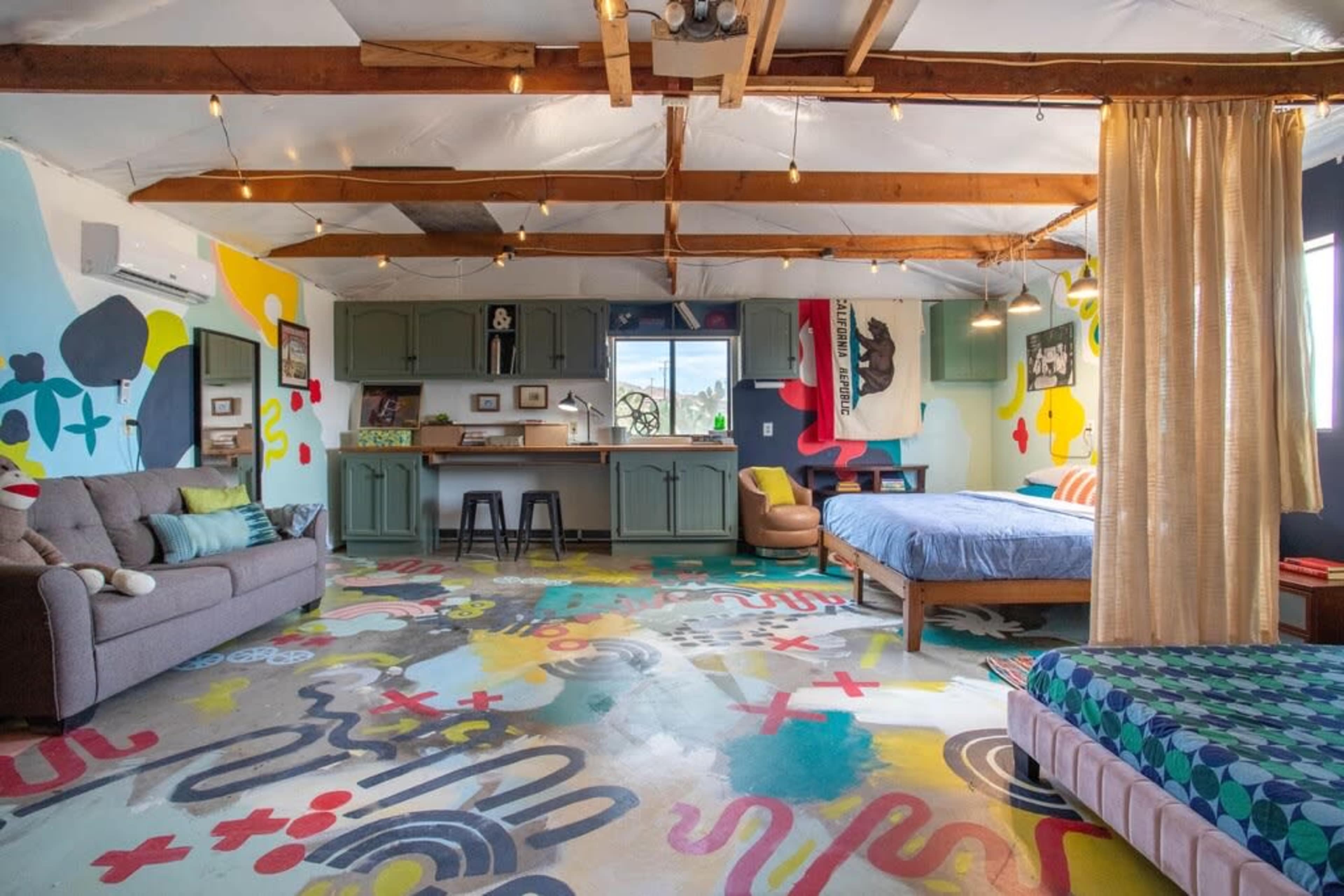 A brightly colored bedroom with a mural-like floor, a sofa, two beds, and a kitchenette area against one wall.