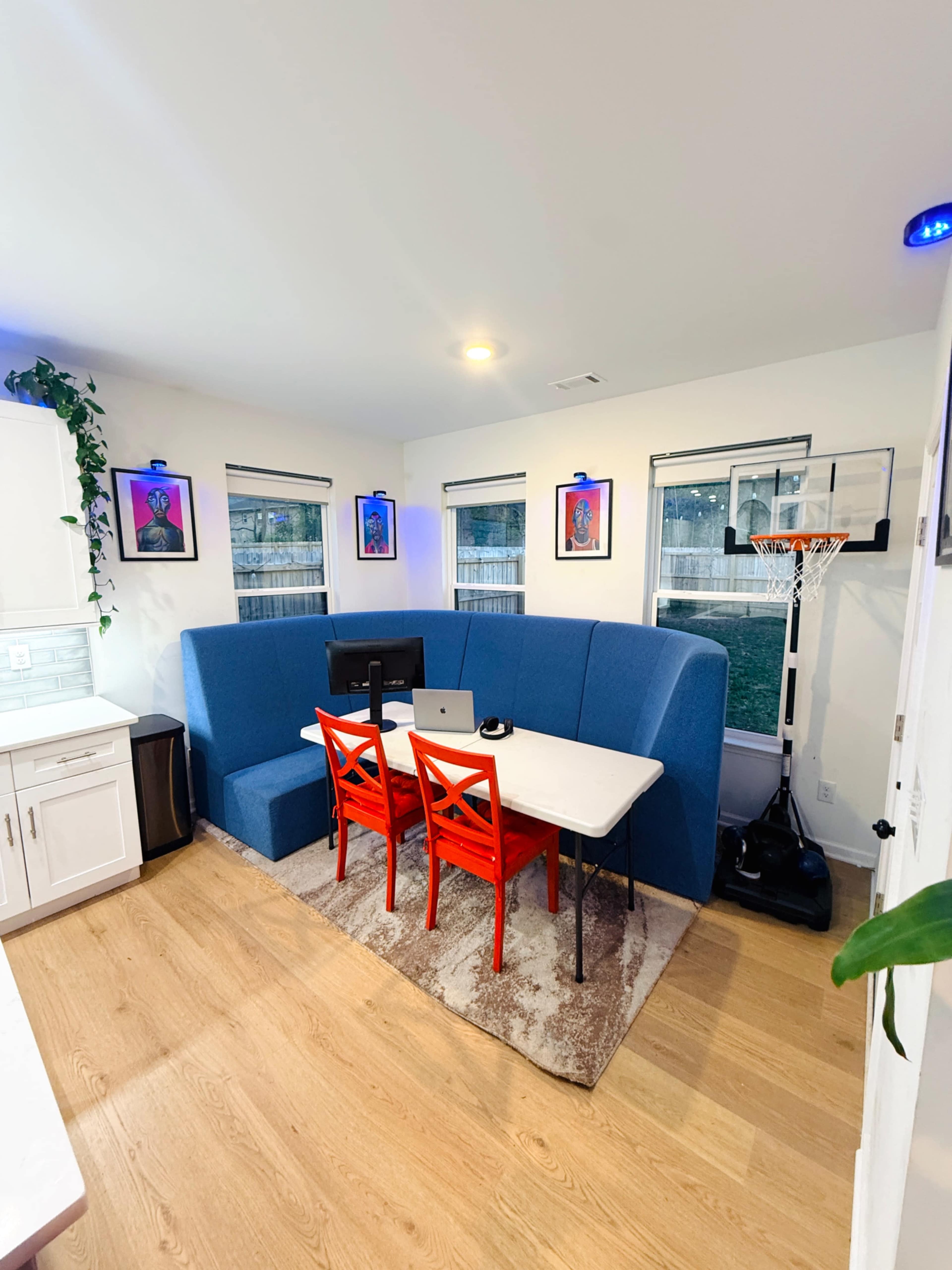 The image shows a small home workspace featuring a blue booth seating area, a white table with a computer, and red chairs, set against a backdrop of windows and framed artwork.