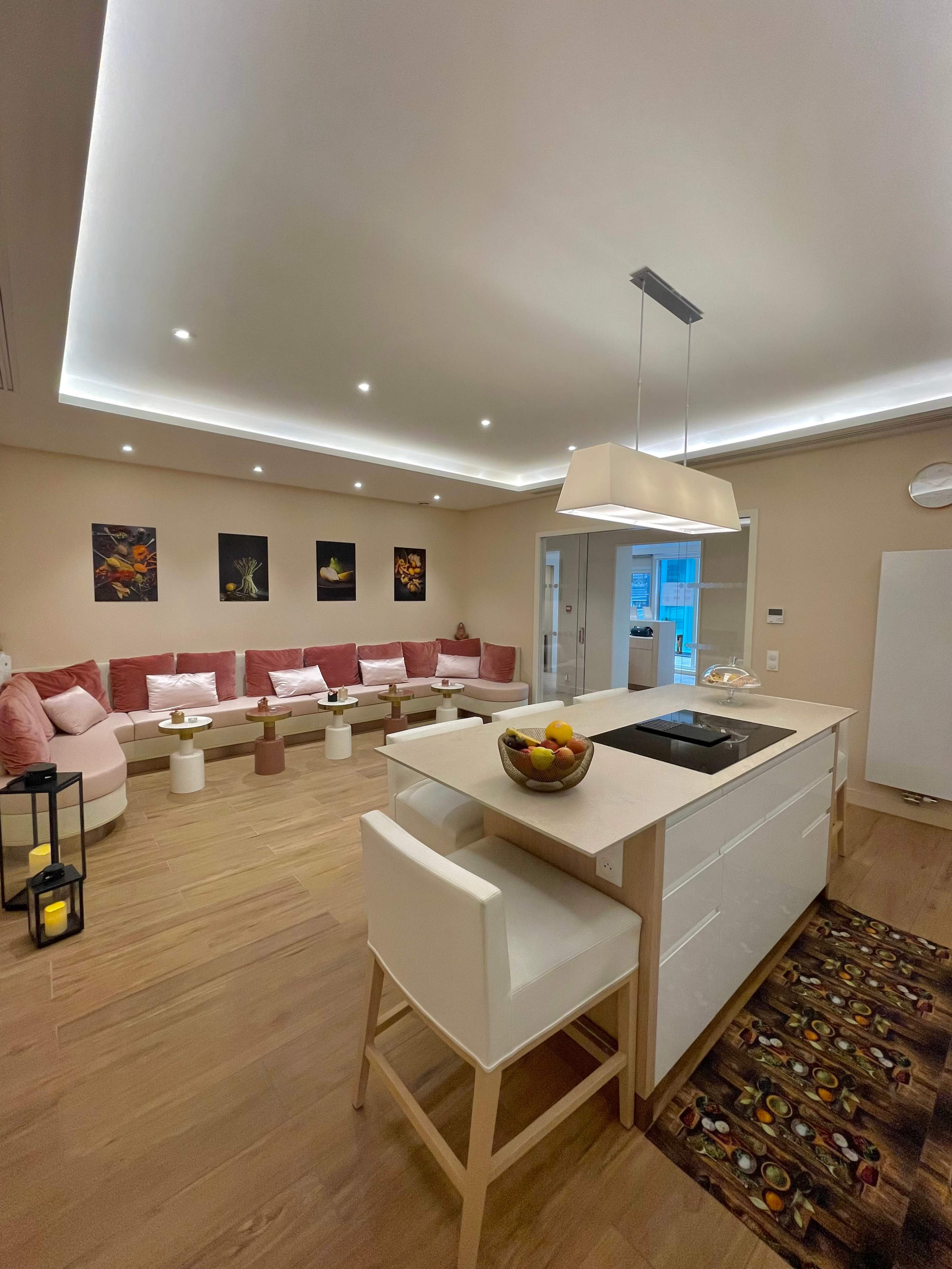 A modern living room features a pink sectional sofa, a sleek kitchen island with a bowl of fruit, and ambient lighting throughout.