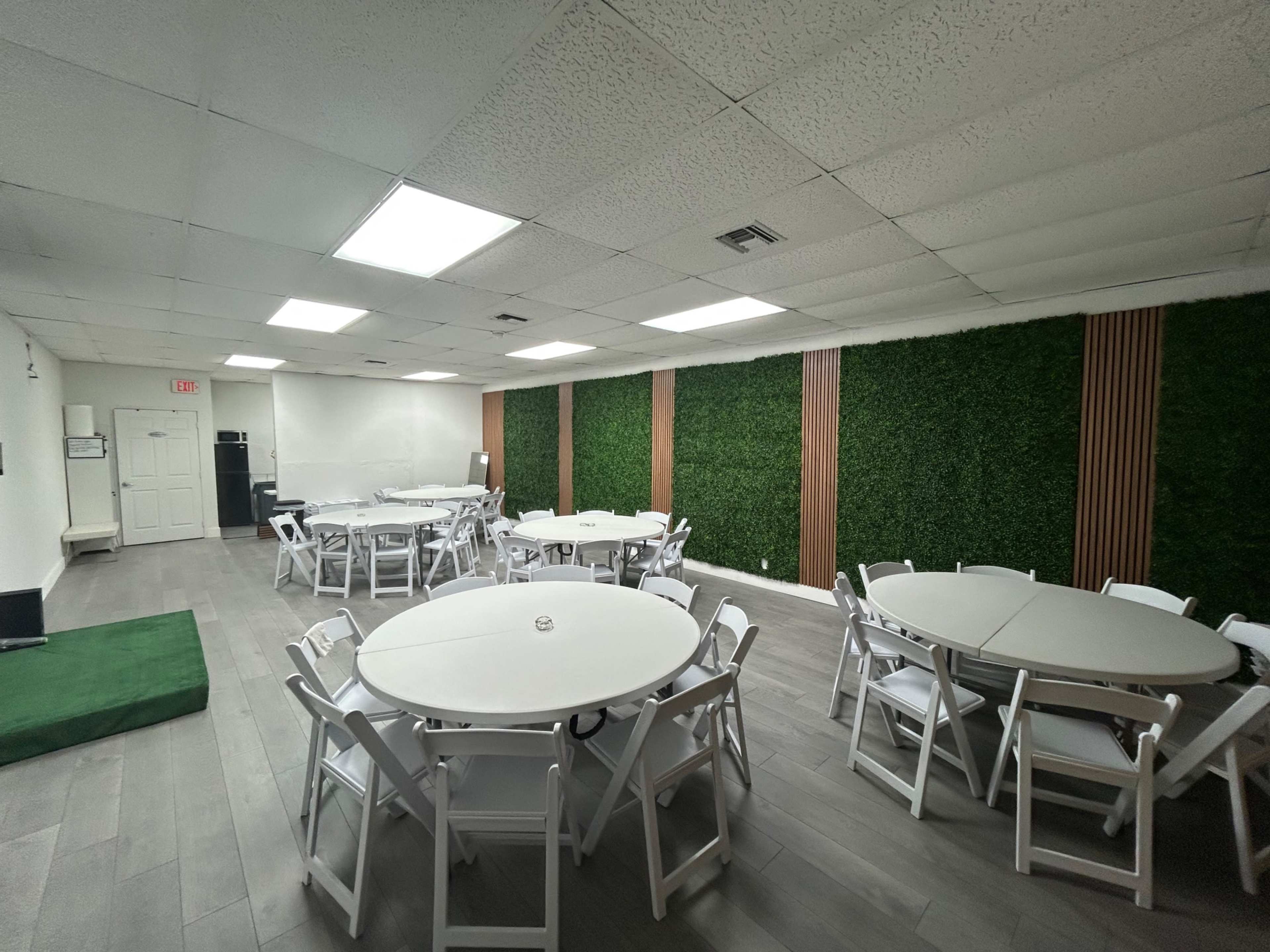 The image shows a spacious room with several round tables and white chairs arranged neatly, alongside a green wall feature on one side.