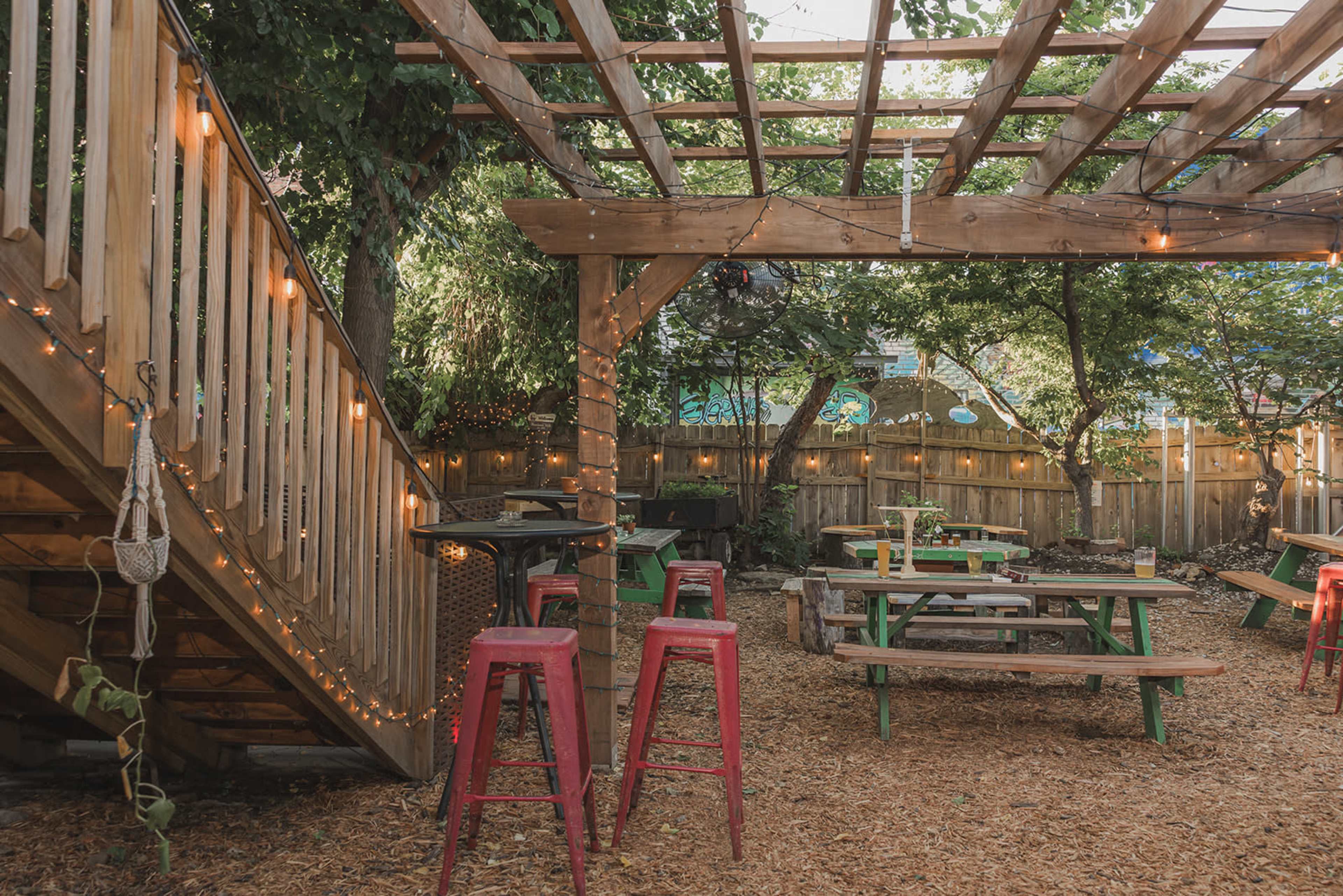 The image showcases an outdoor patio area with wooden tables and chairs, surrounded by greenery and string lights overhead.