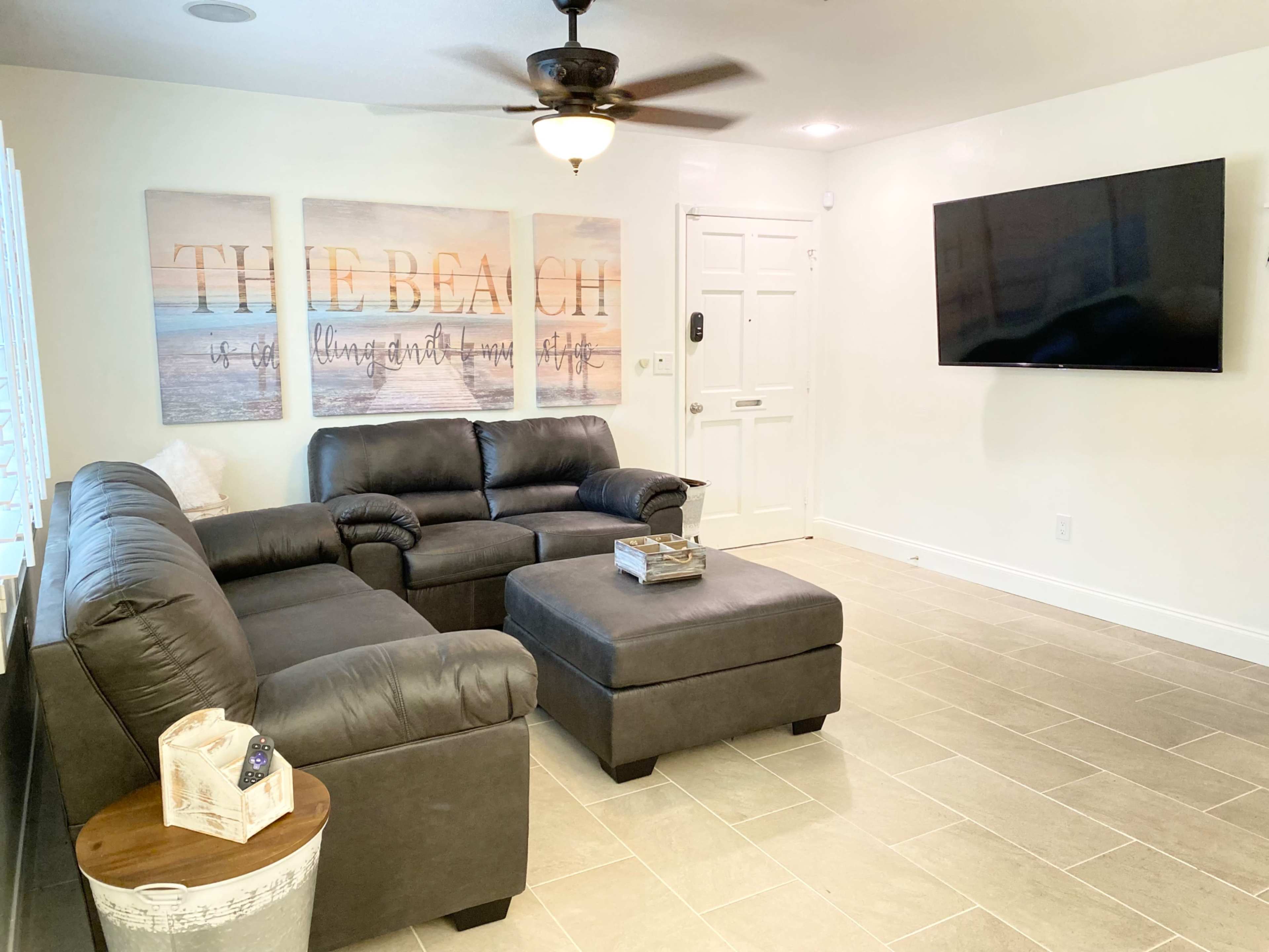 A living room featuring a dark leather sectional sofa, a matching ottoman, a television mounted on the wall, and three framed beach-themed artworks above the couch.