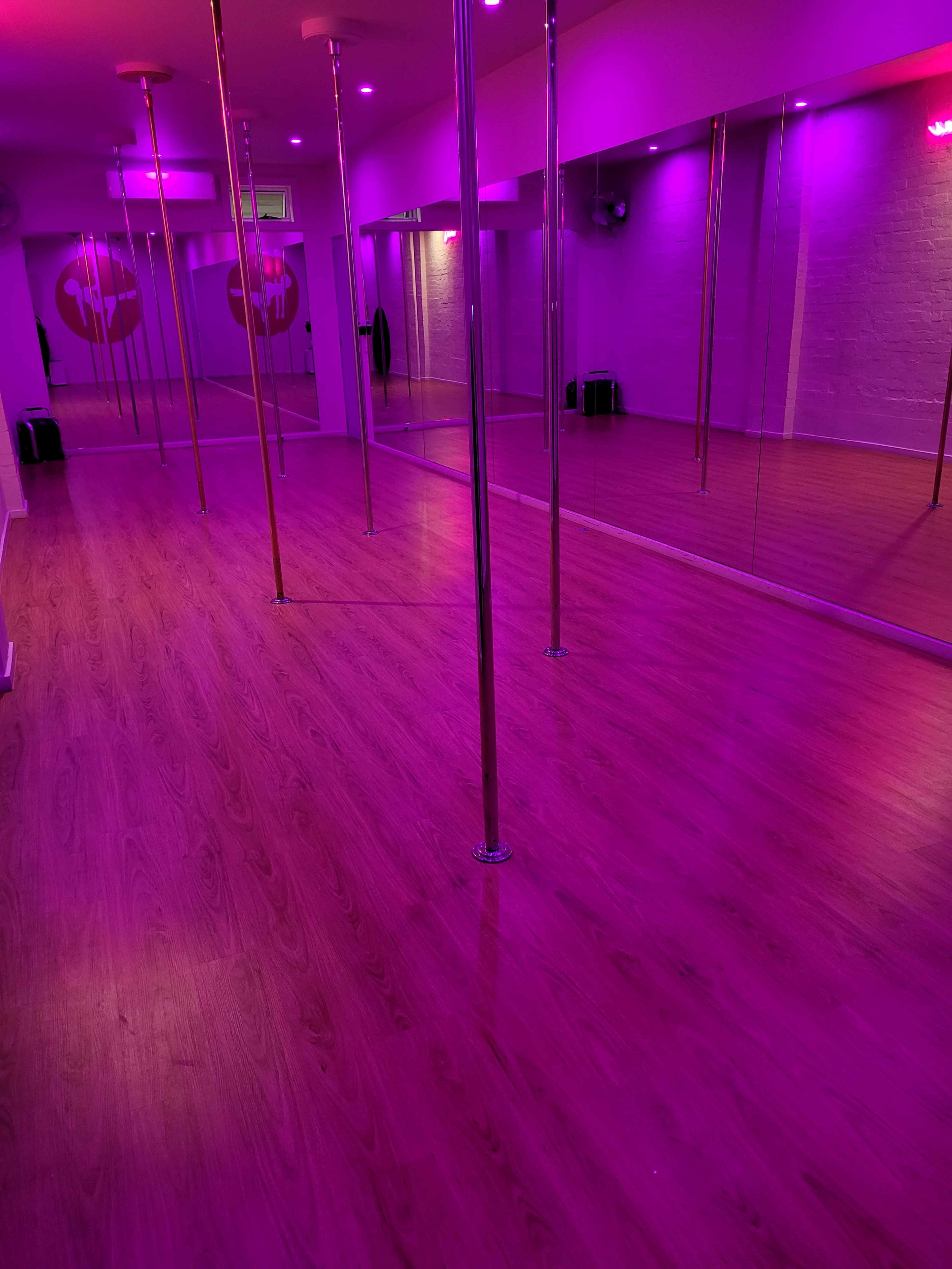 A dance studio with mirrored walls, purple lighting, and several vertical poles set against a wooden floor.
