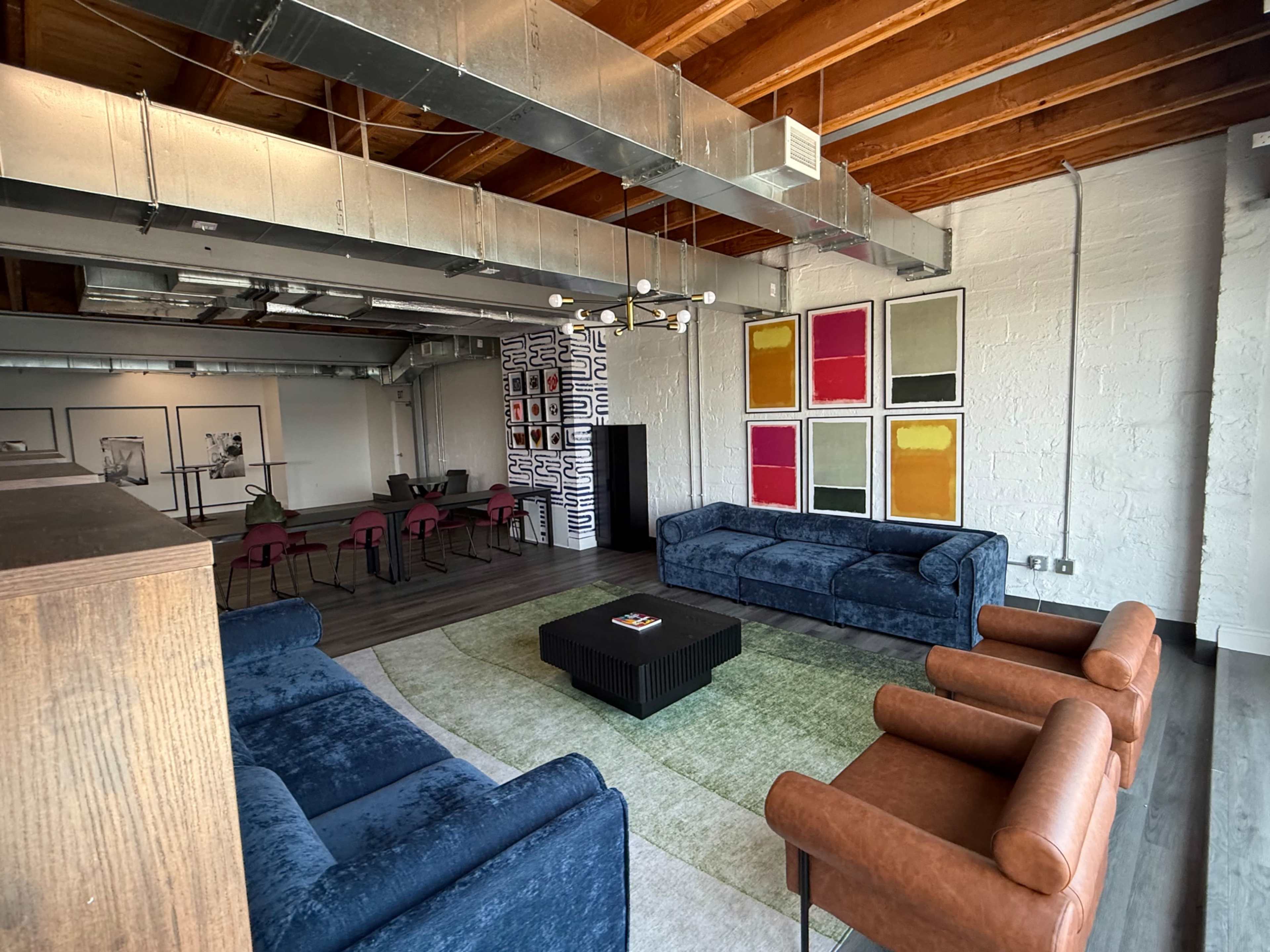 The image shows a modern room with blue sofas, brown chairs, a black coffee table, and a colorful wall featuring four framed art pieces.