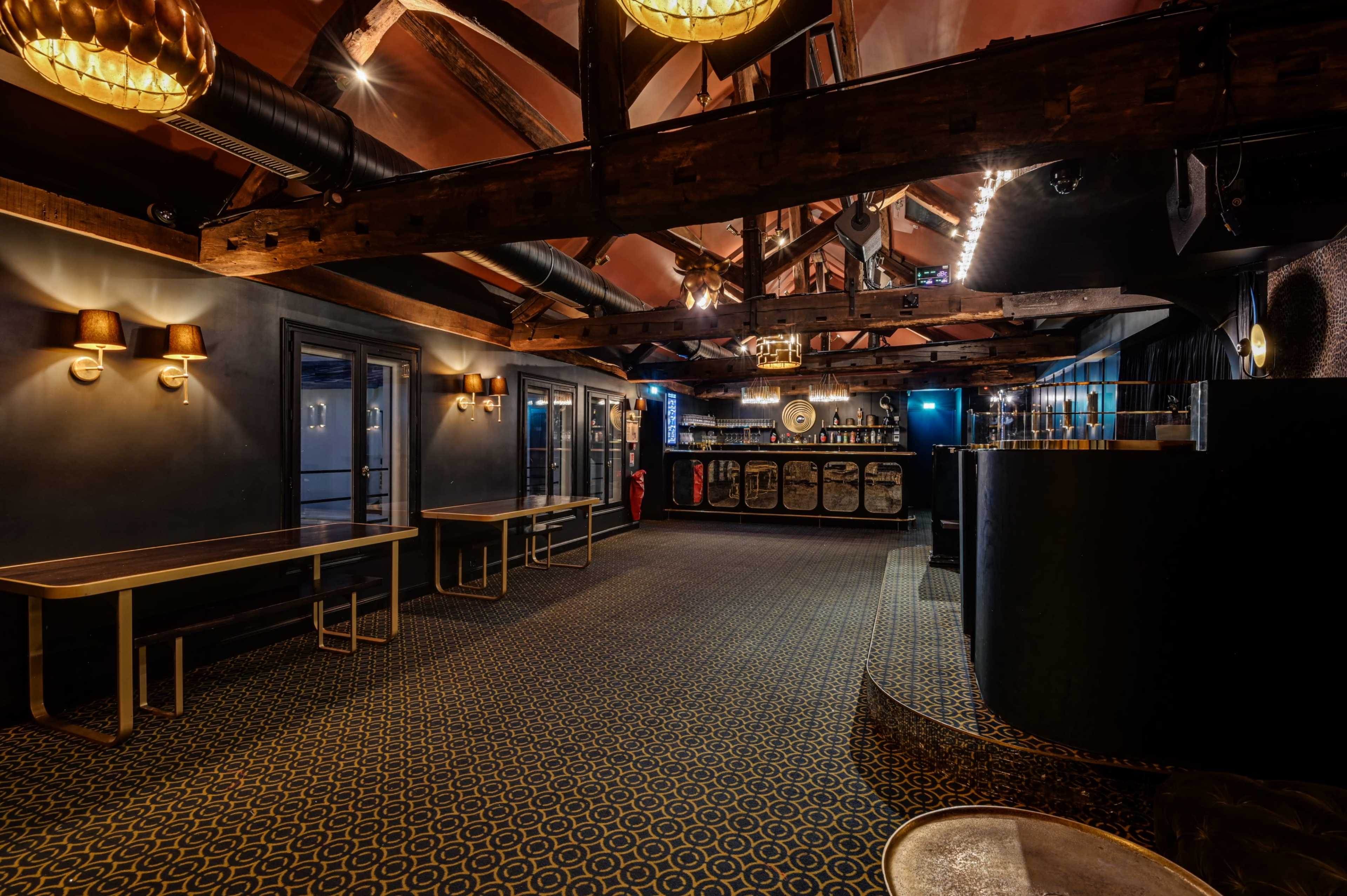 The image shows a spacious interior of a bar with wooden beams, low-light fixtures, and a bar counter in the background.