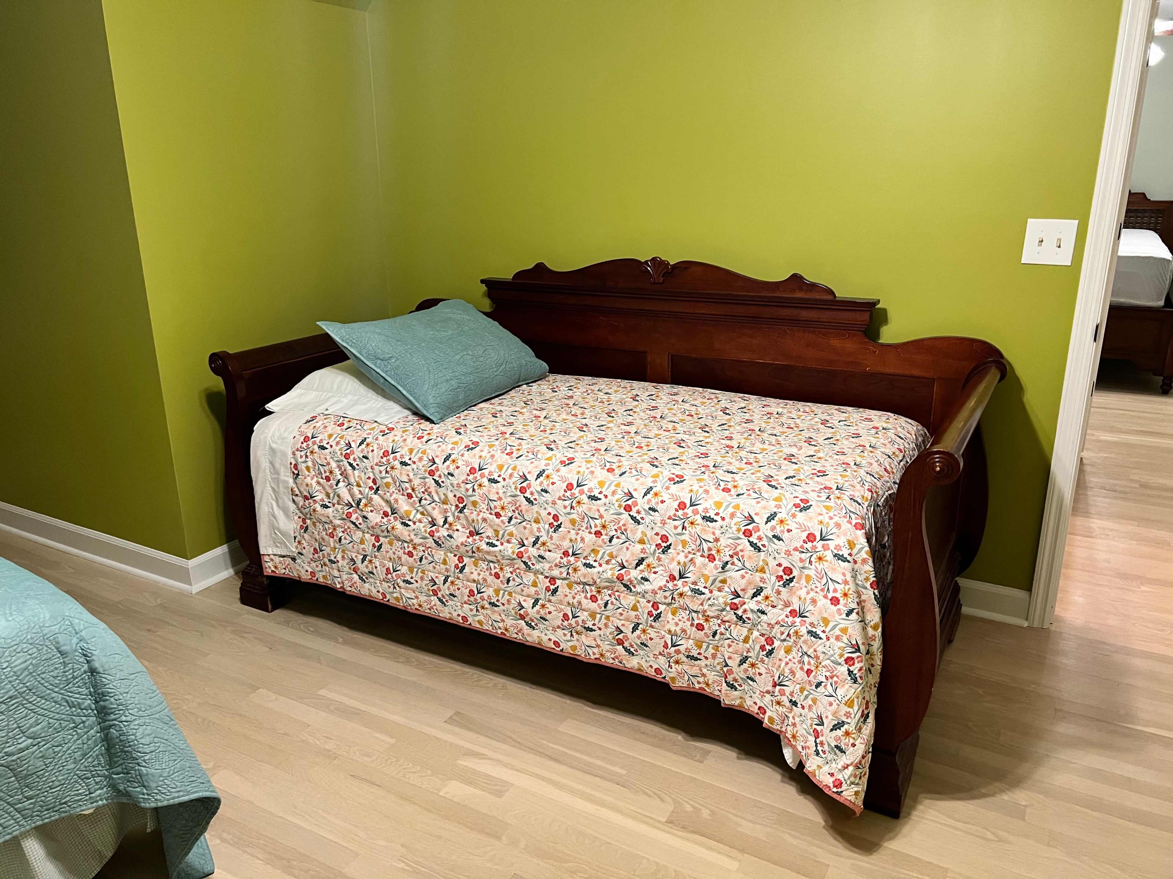 The image shows a wooden daybed with a floral-patterned blanket and a light blue pillow against a green wall.