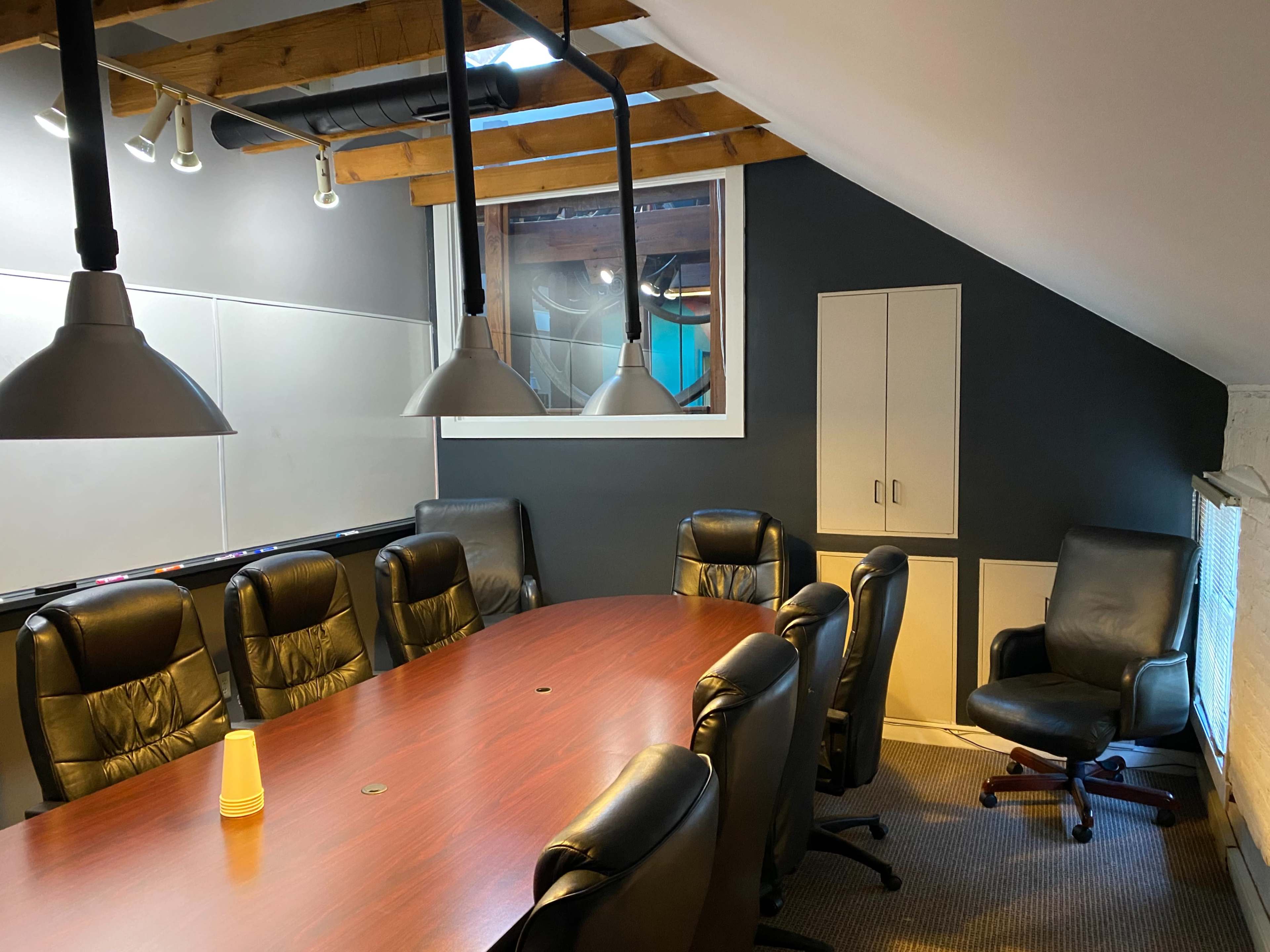 The image shows a conference room with a long wooden table, black leather chairs, and pendant lights hanging from a sloped ceiling.