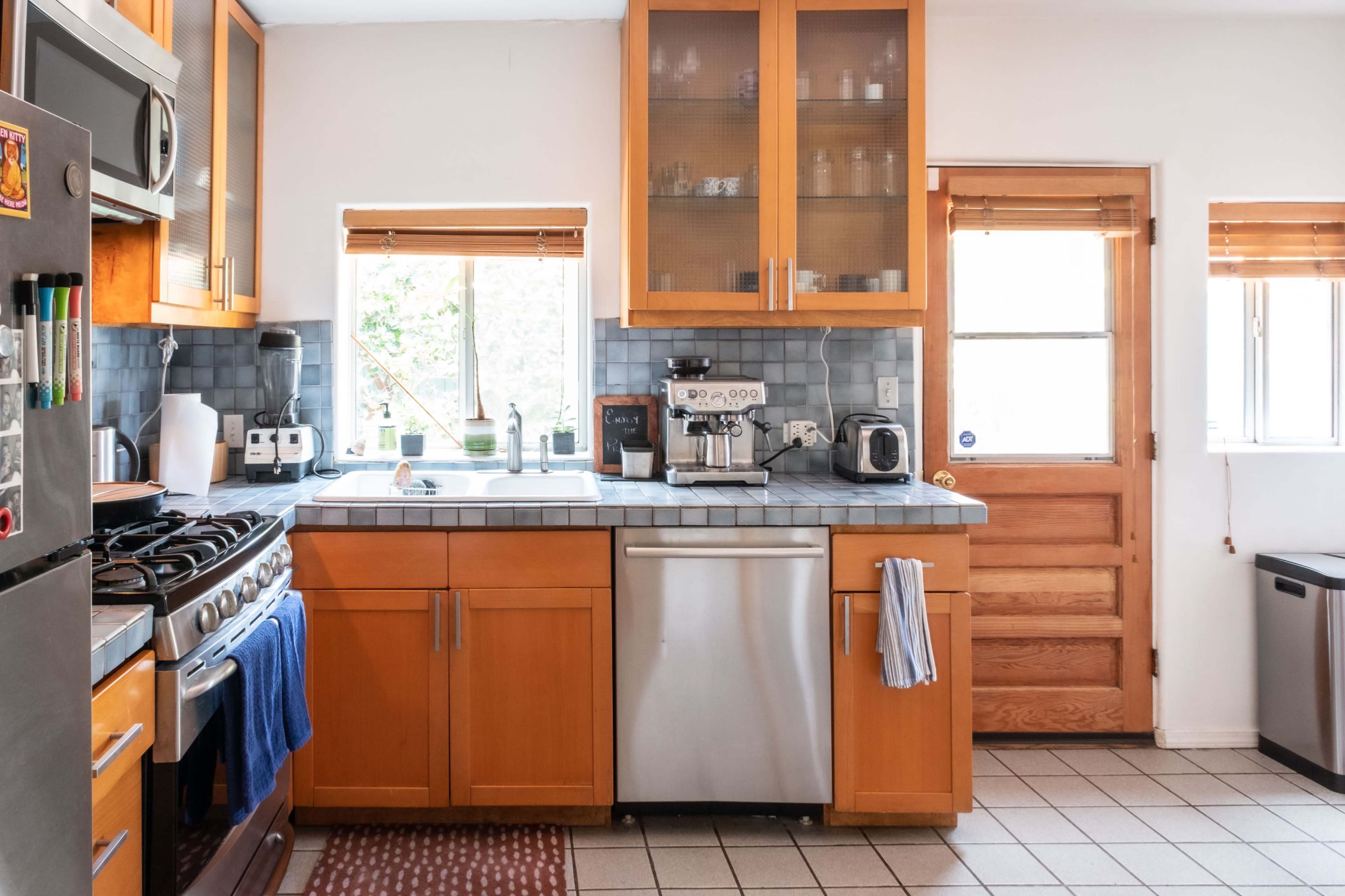 The image shows a modern kitchen with wooden cabinets, a stainless steel dishwasher, and a gas stove, featuring a window that allows natural light and a wooden door leading outside.