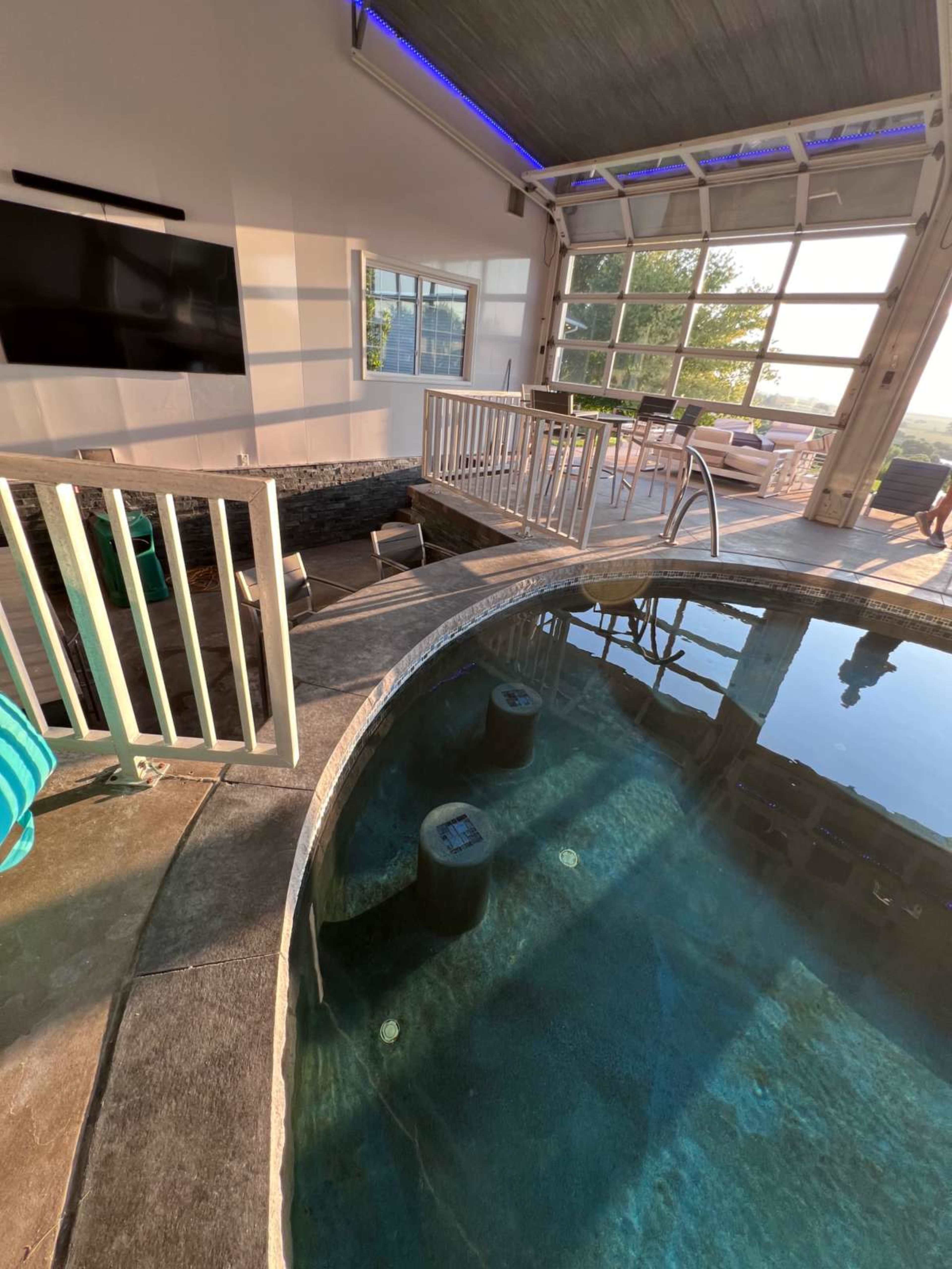 The image shows a modern indoor-outdoor pool area with a circular pool, a television mounted on the wall, and seating nearby, all enclosed by large glass doors.