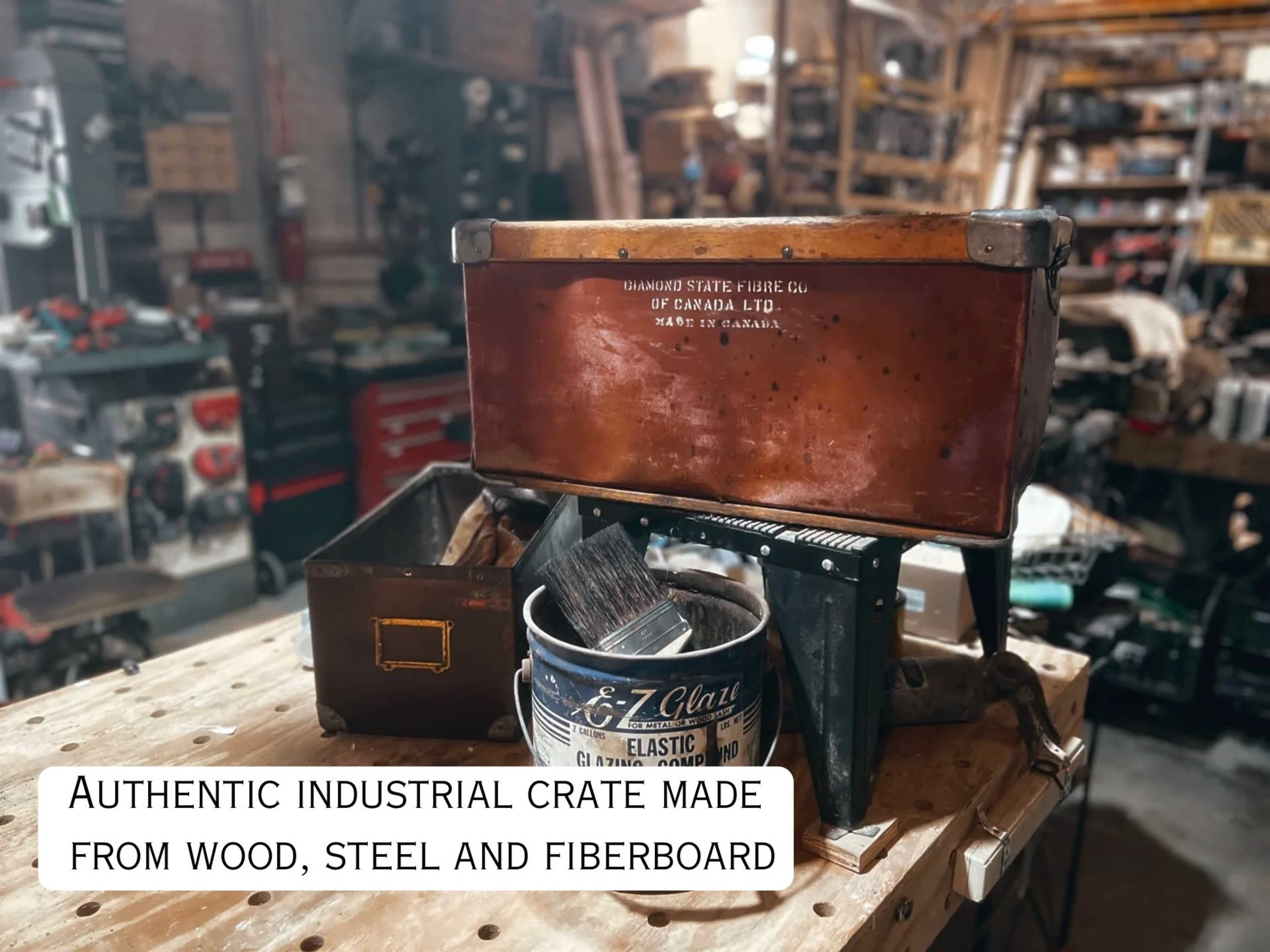 The image shows an authentic industrial crate made from wood, steel, and fiberboard placed on a workbench in a workshop filled with various tools and equipment.