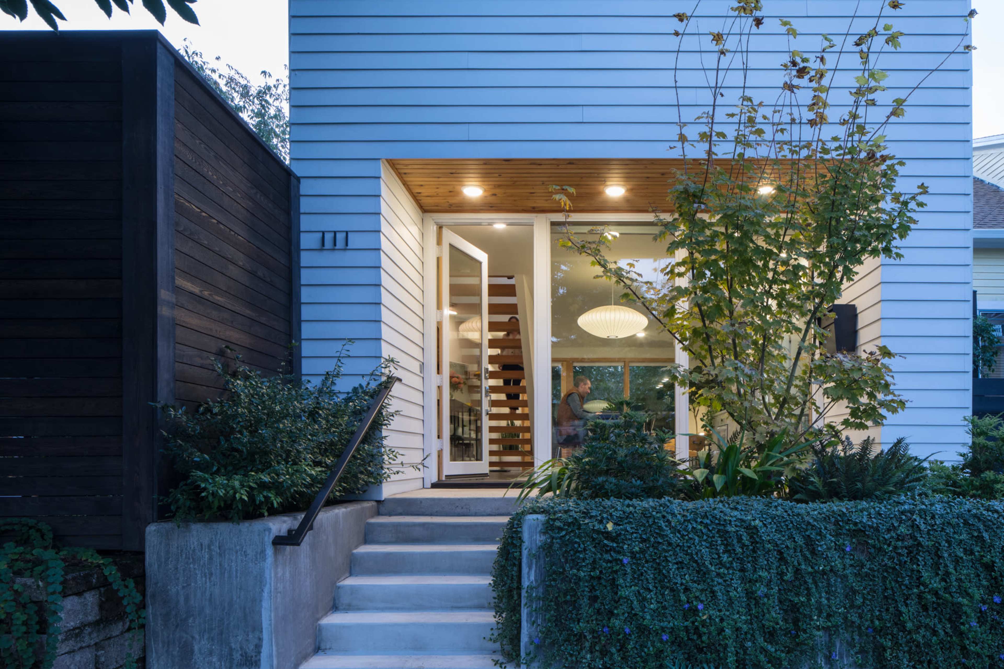 A modern house features a front entrance with a stepped walkway, surrounded by greenery and natural light illuminating the interior.