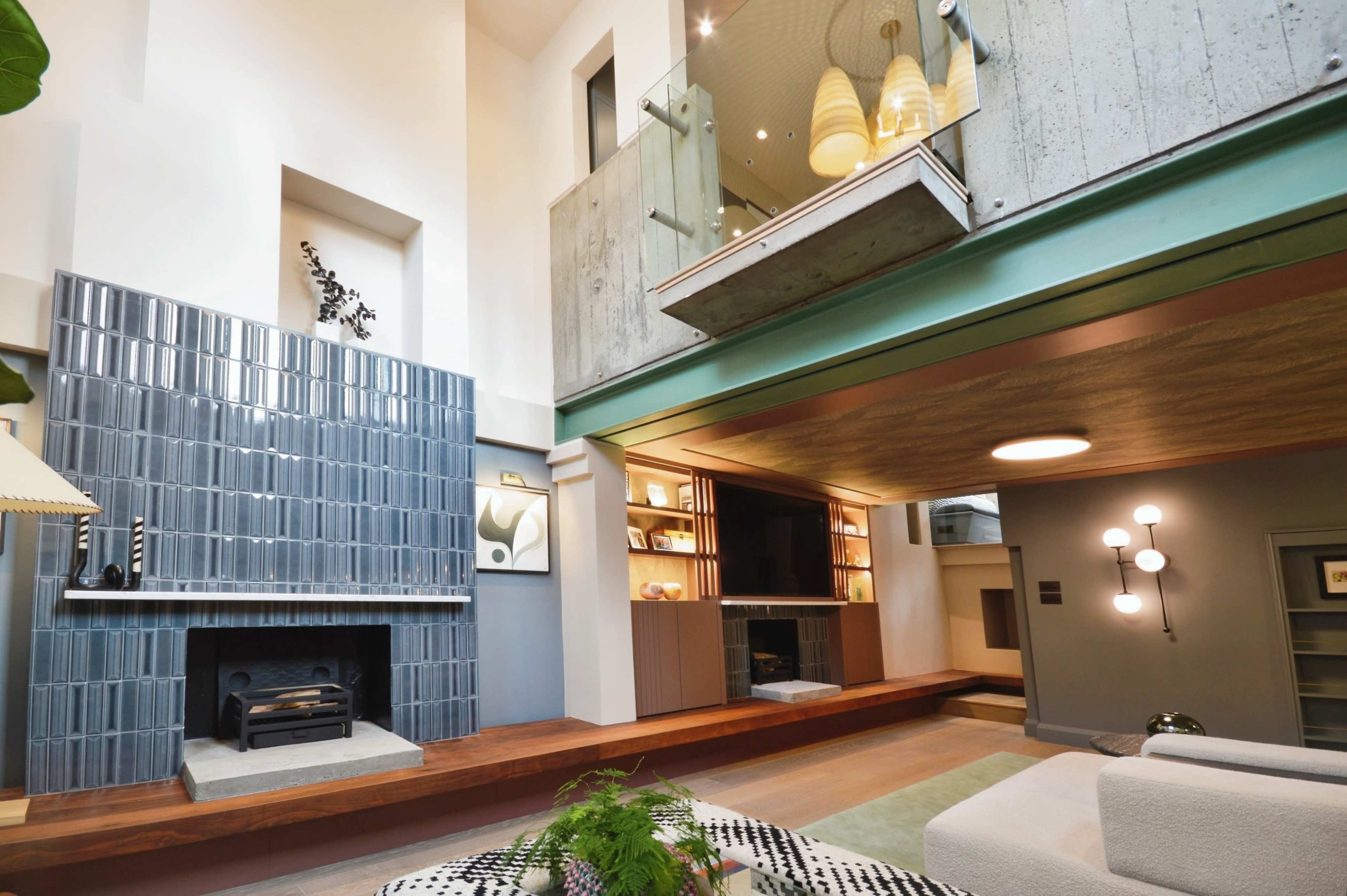 The image shows a modern living room with a linear fireplace, a large glass balcony above, and a combination of textured walls and wooden flooring.