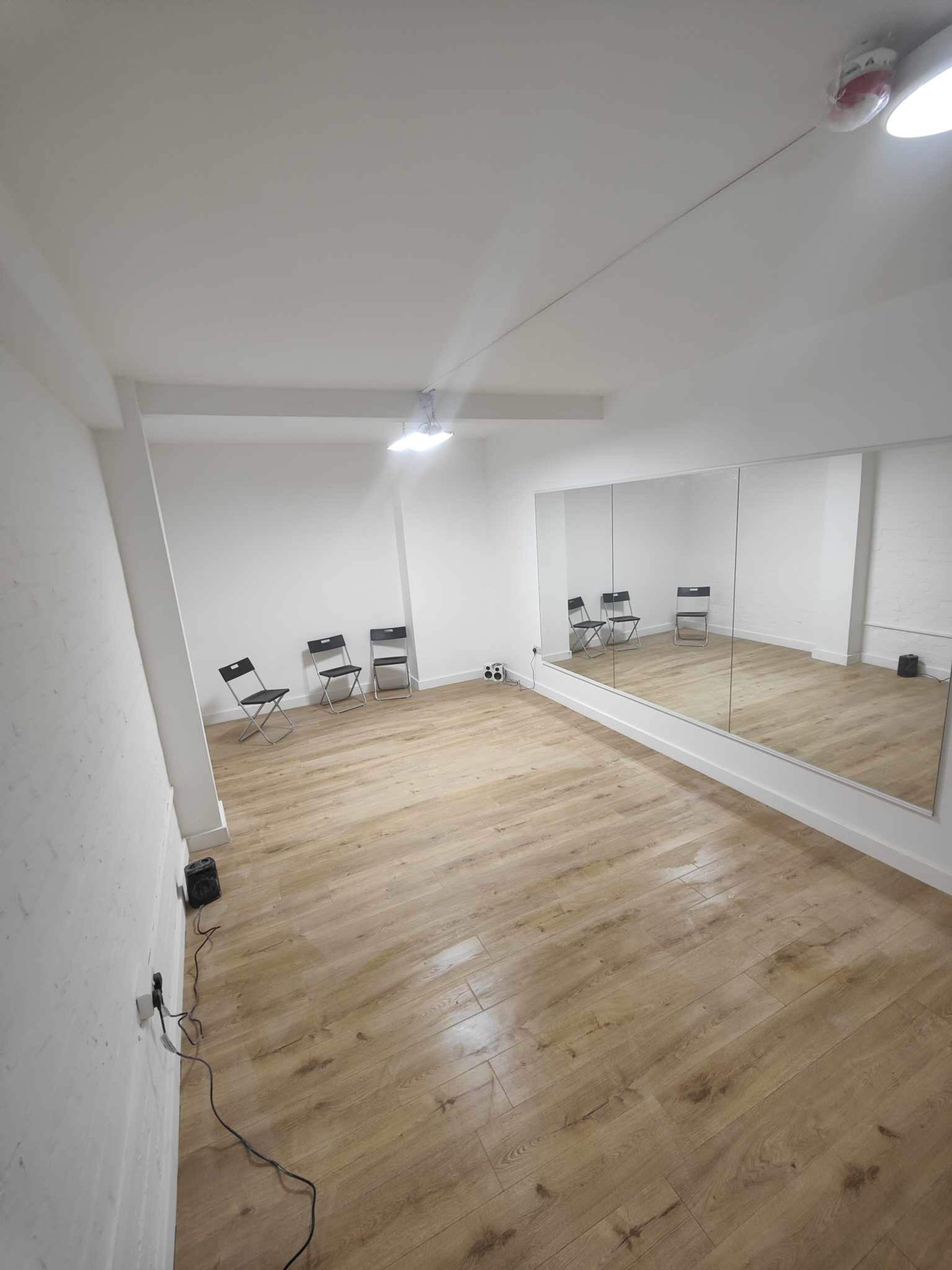 The image shows a minimalist room with wooden flooring, a mirrored wall, and several folding chairs positioned against one side.