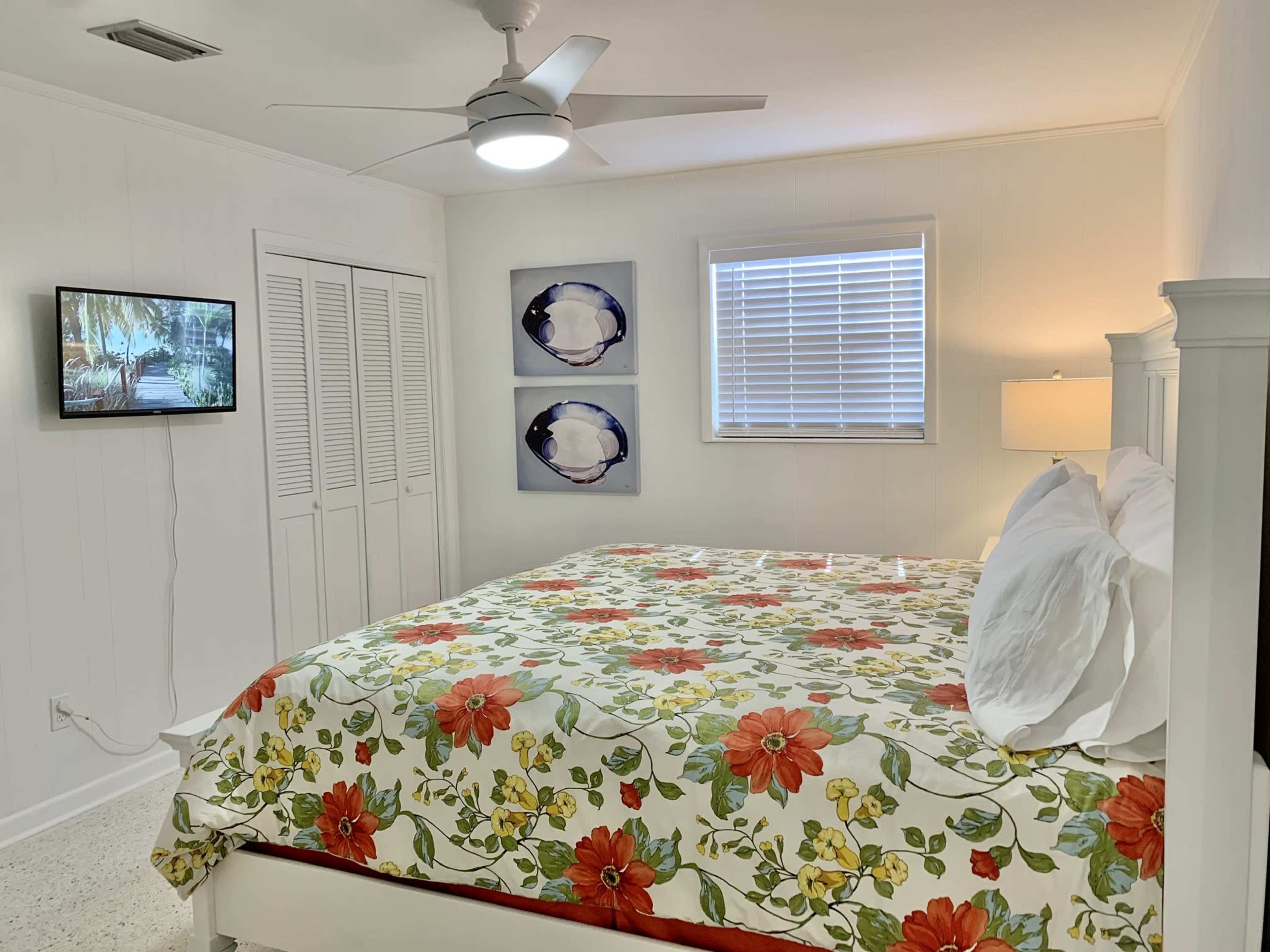 A bright bedroom with a large bed covered in a floral quilt, a mounted TV on the wall, and two framed pictures above the bed.