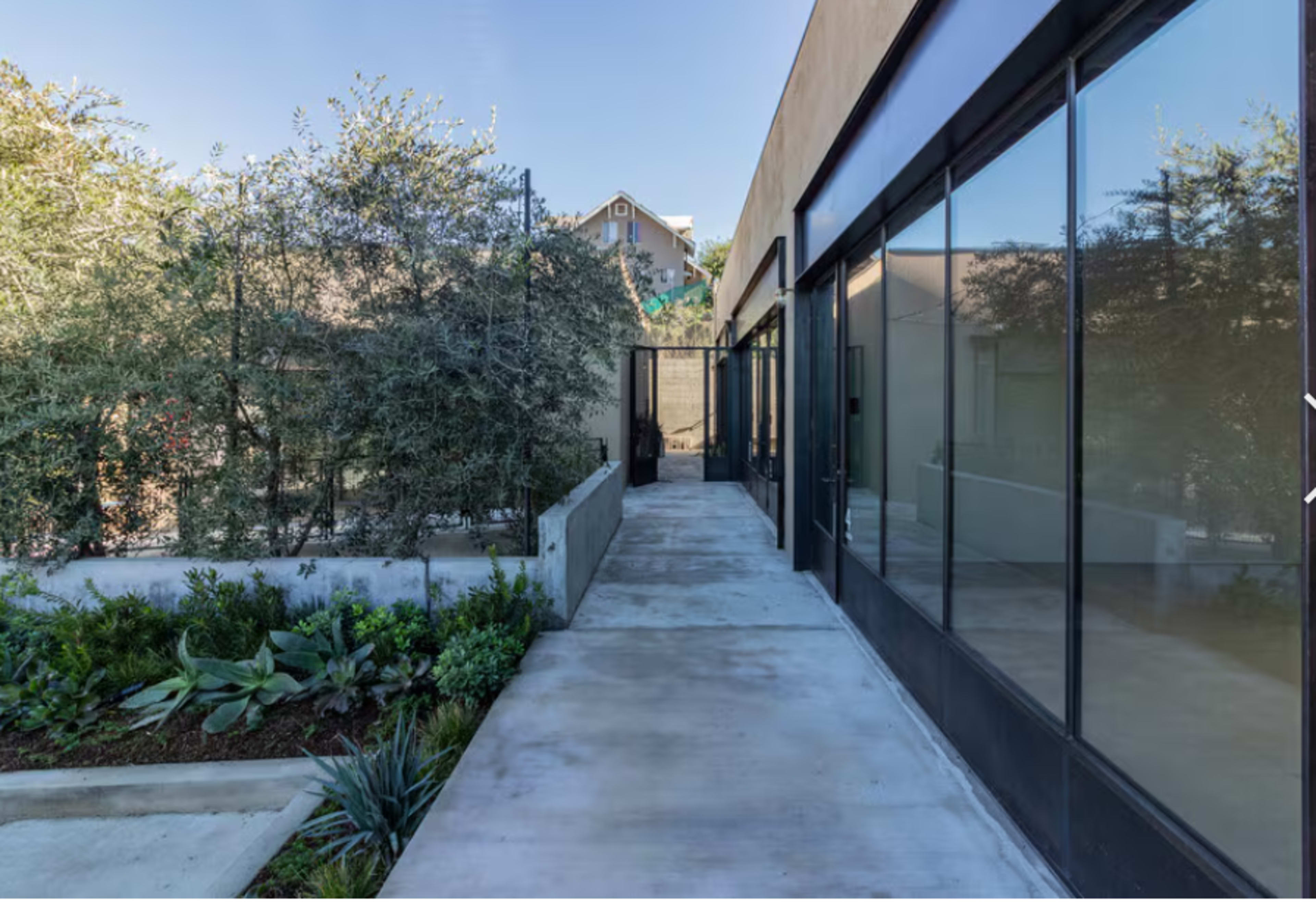 The image shows a modern, narrow pathway leading between two buildings, with glass walls on one side and greenery along the other.