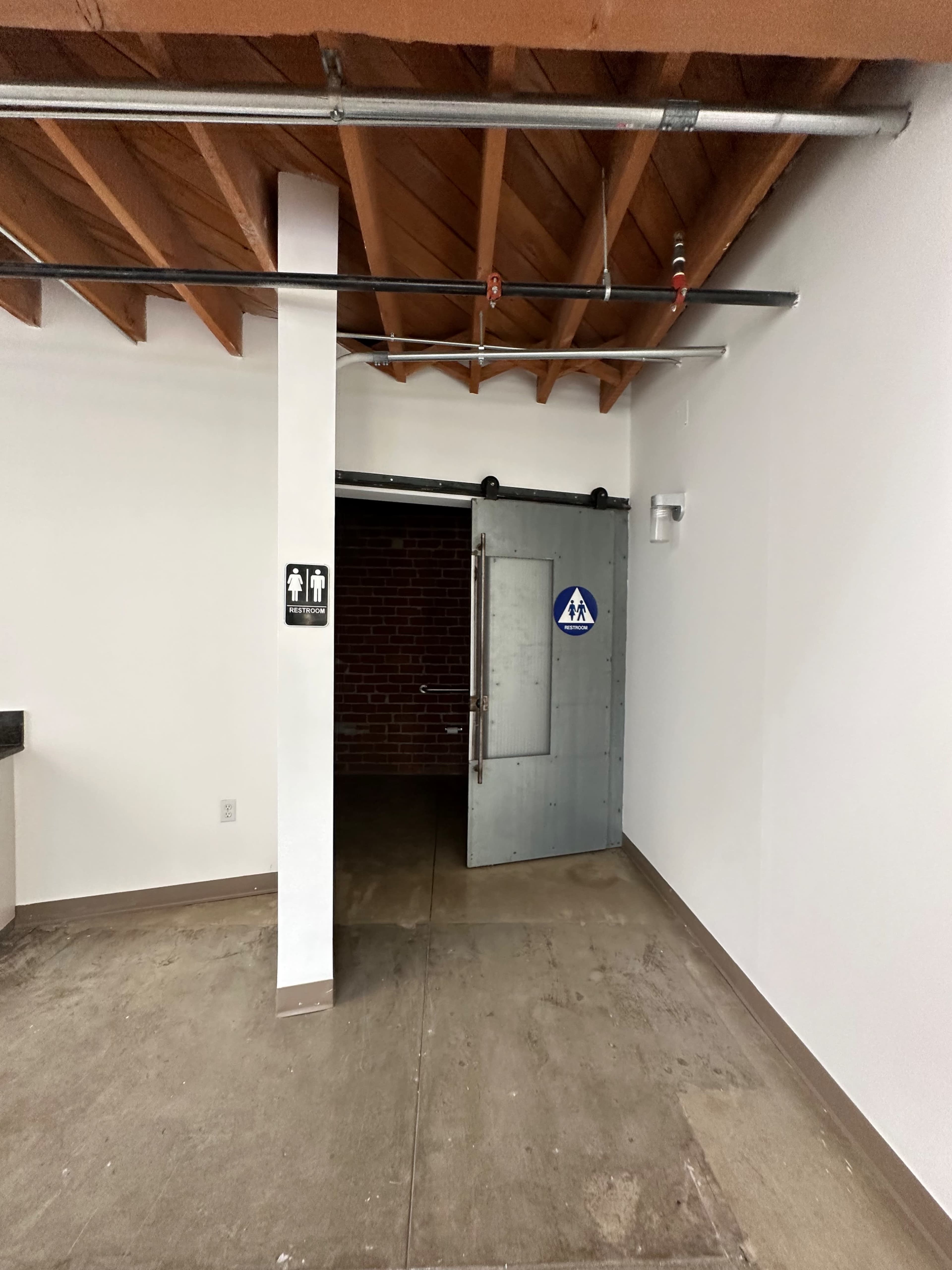 The image shows an interior space featuring a door marked with restroom signs, leading to a dimly lit area with exposed beams and concrete flooring.