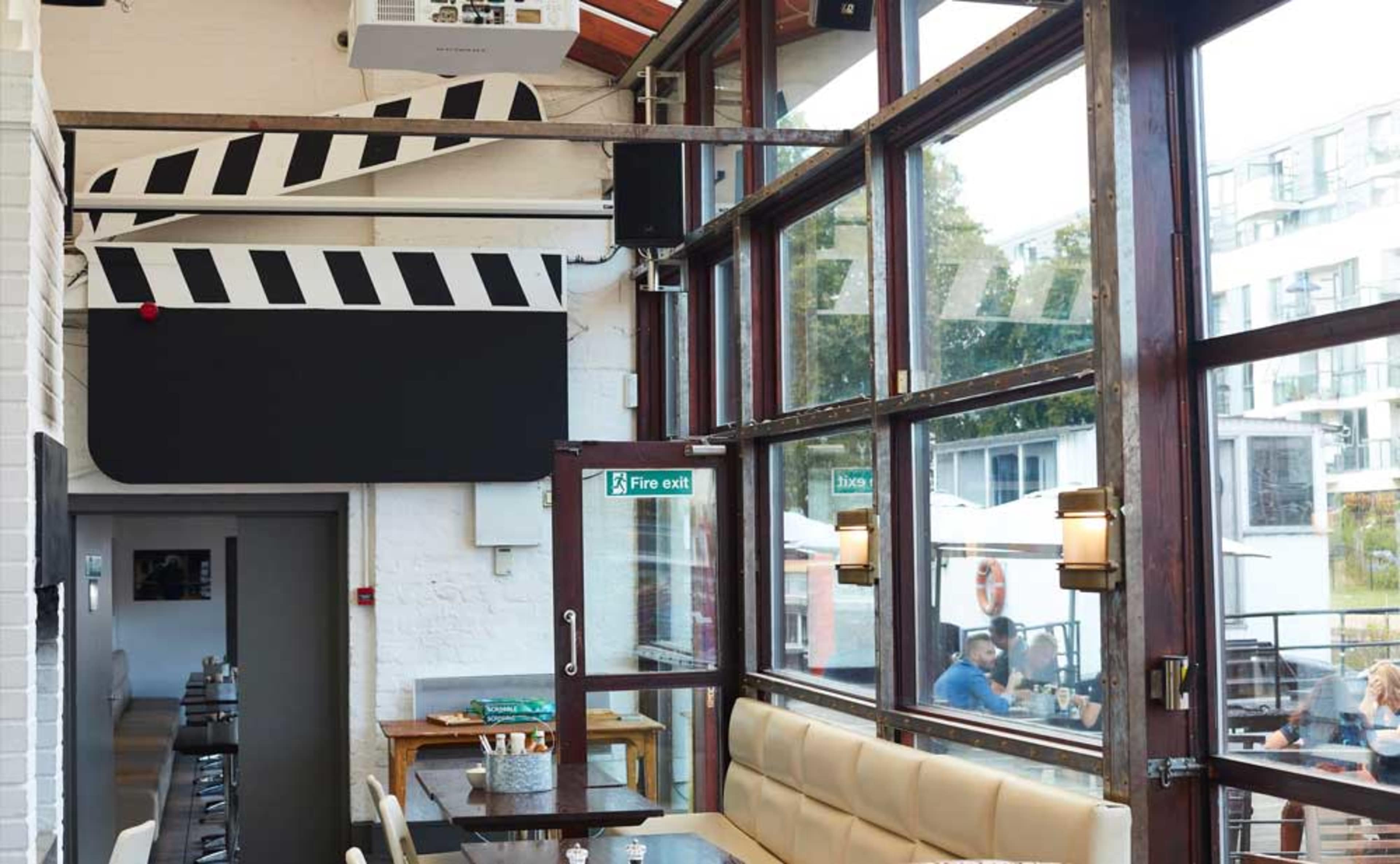 The interior of a restaurant features large windows, wooden furnishings, and a film clapperboard motif on the wall.