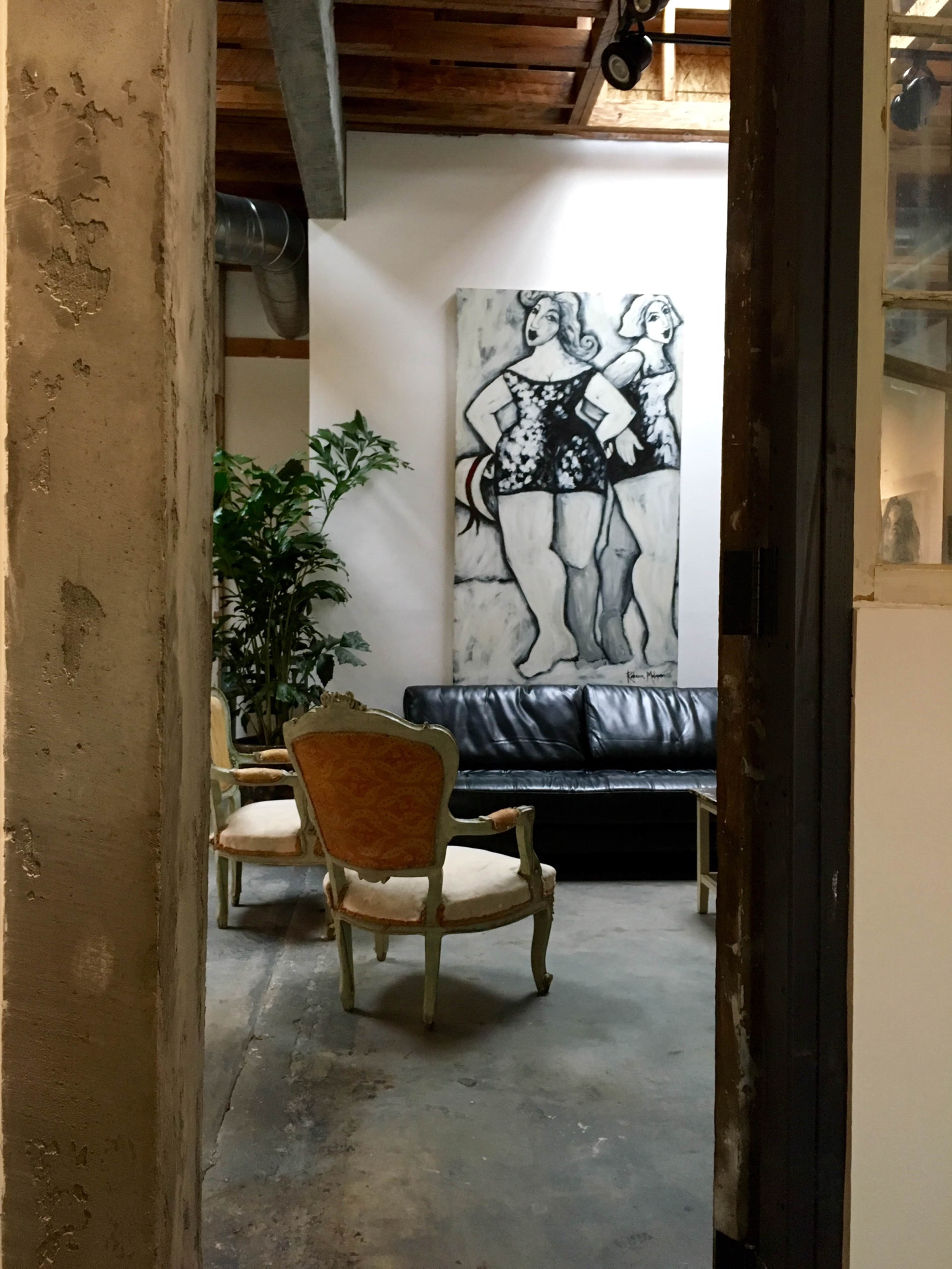 A hallway leads to a sitting area with a black leather couch, two chairs, a large artwork on the wall, and a potted plant.
