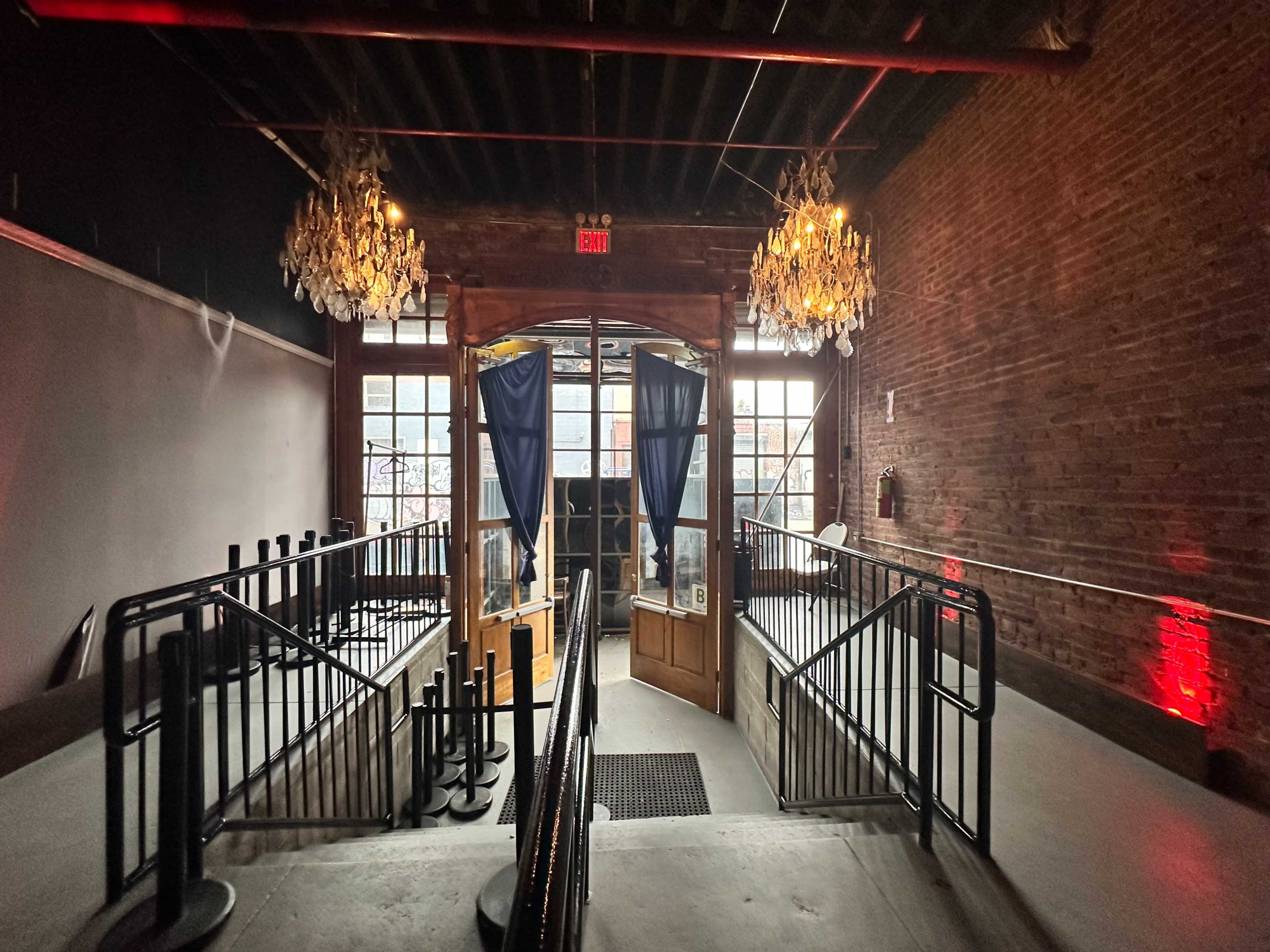 An interior space featuring an entrance with double doors, flanked by two chandeliers, and a staircase with black railings leading down to a partially visible area.