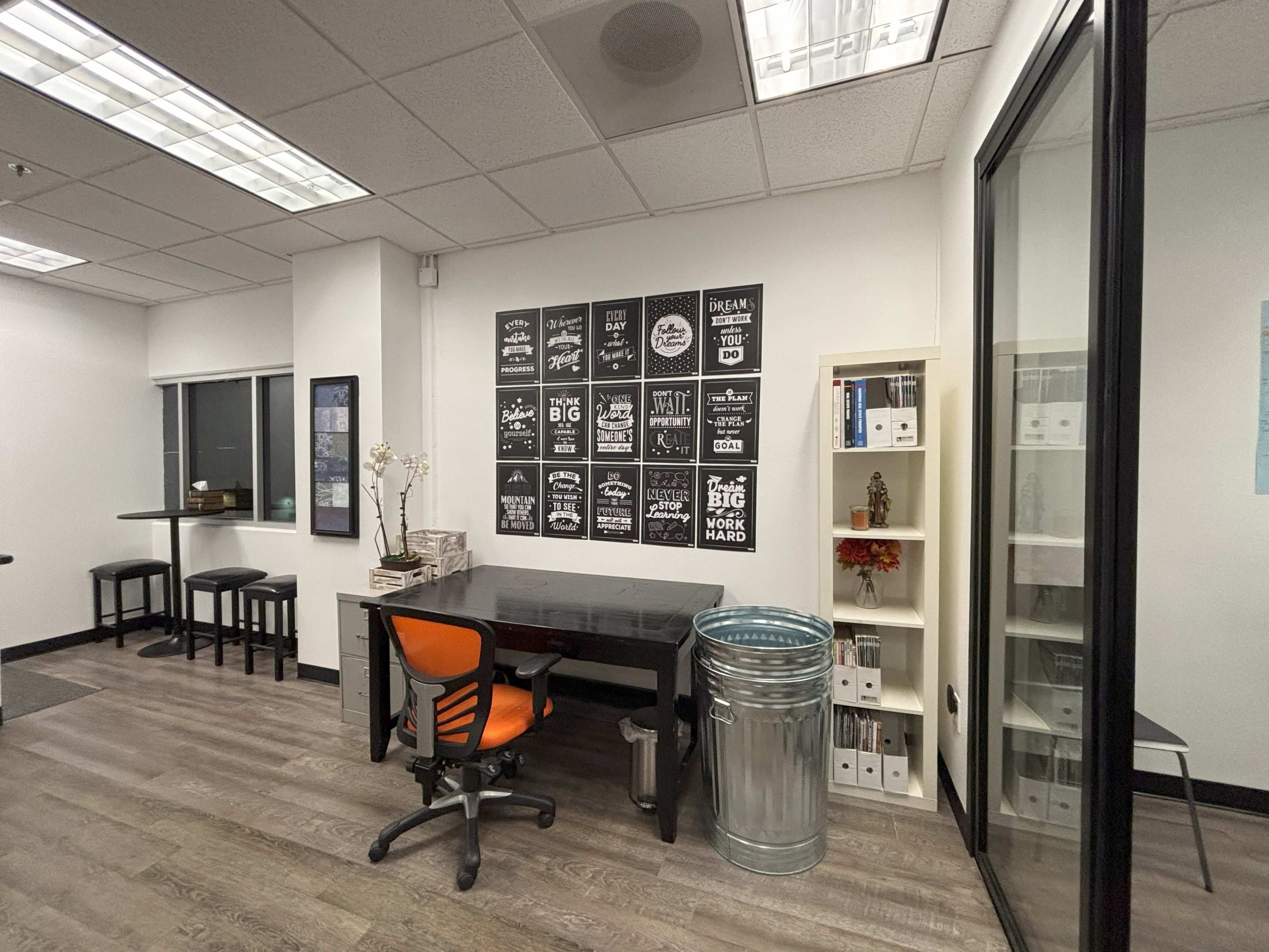 The image shows an office space with a desk and chair, a large trash can, a shelving unit with binders, and motivational posters on the wall.