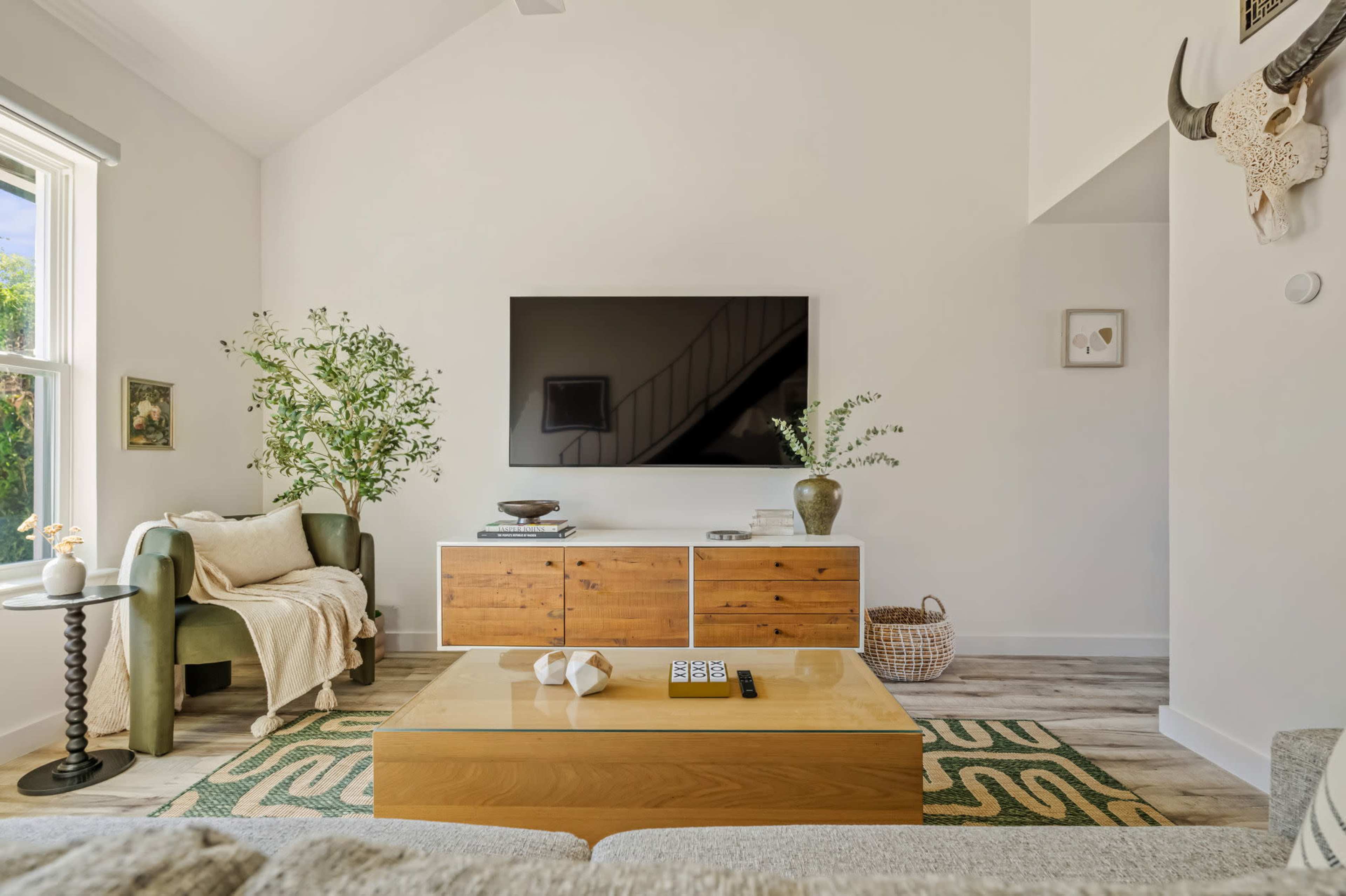 The image shows a bright living room with a large wall-mounted TV, a wooden coffee table, a green chair, and decorative plants.