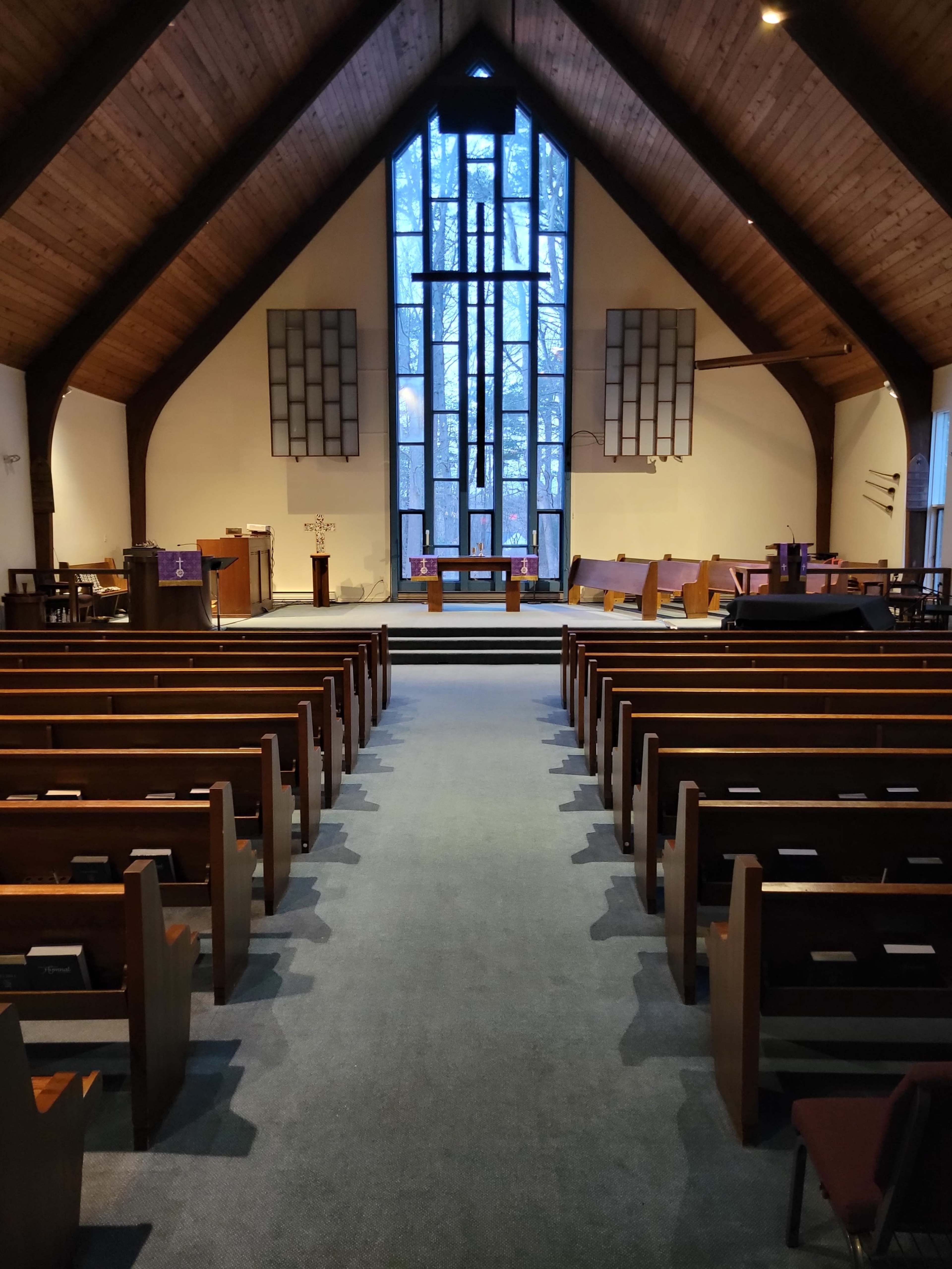 The interior of a church features wooden beams, rows of pews, and a large stained glass window at the front.