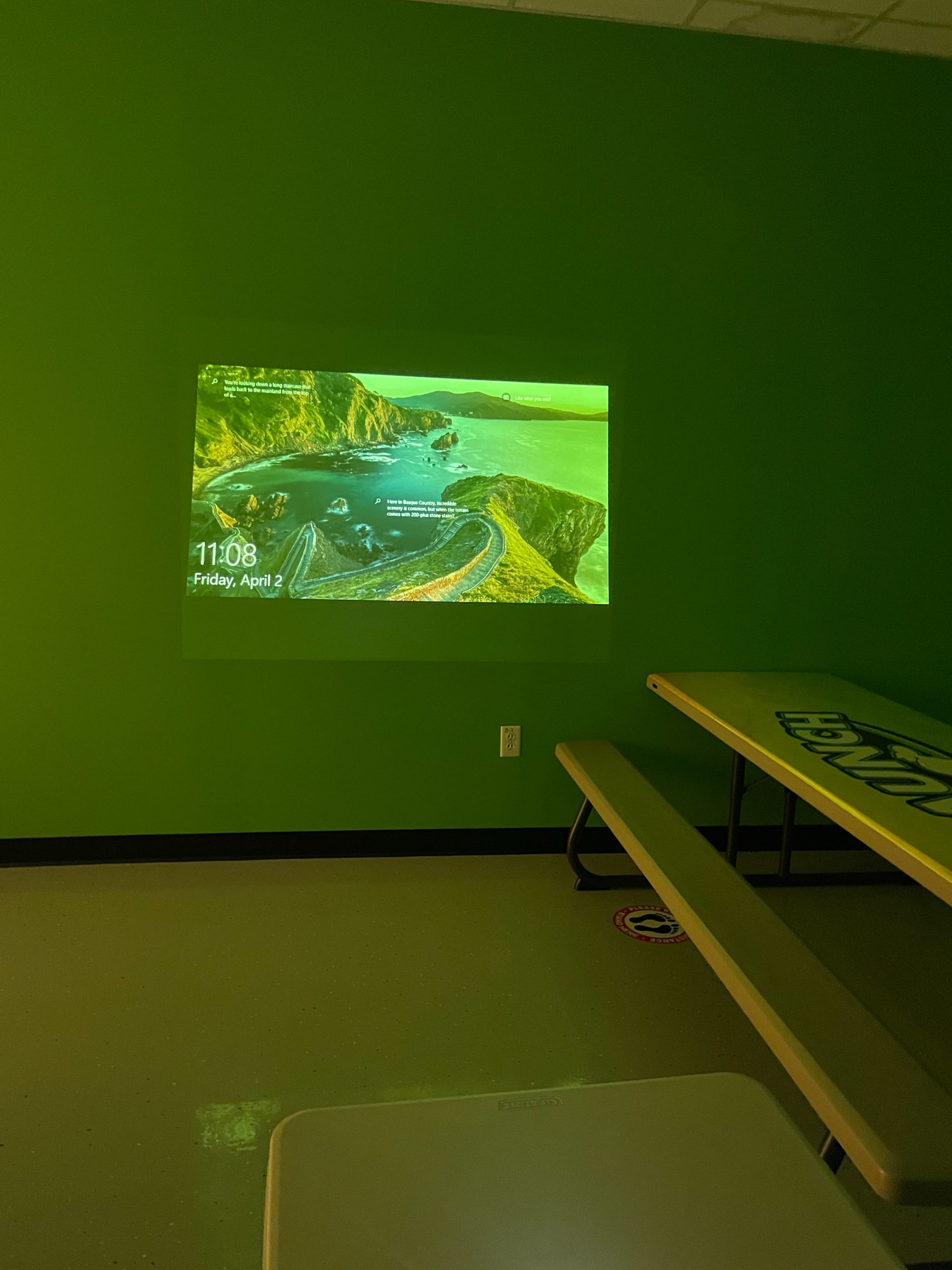 The image shows a green-walled room with a projection of a scenic landscape displaying the time and date on a wall.