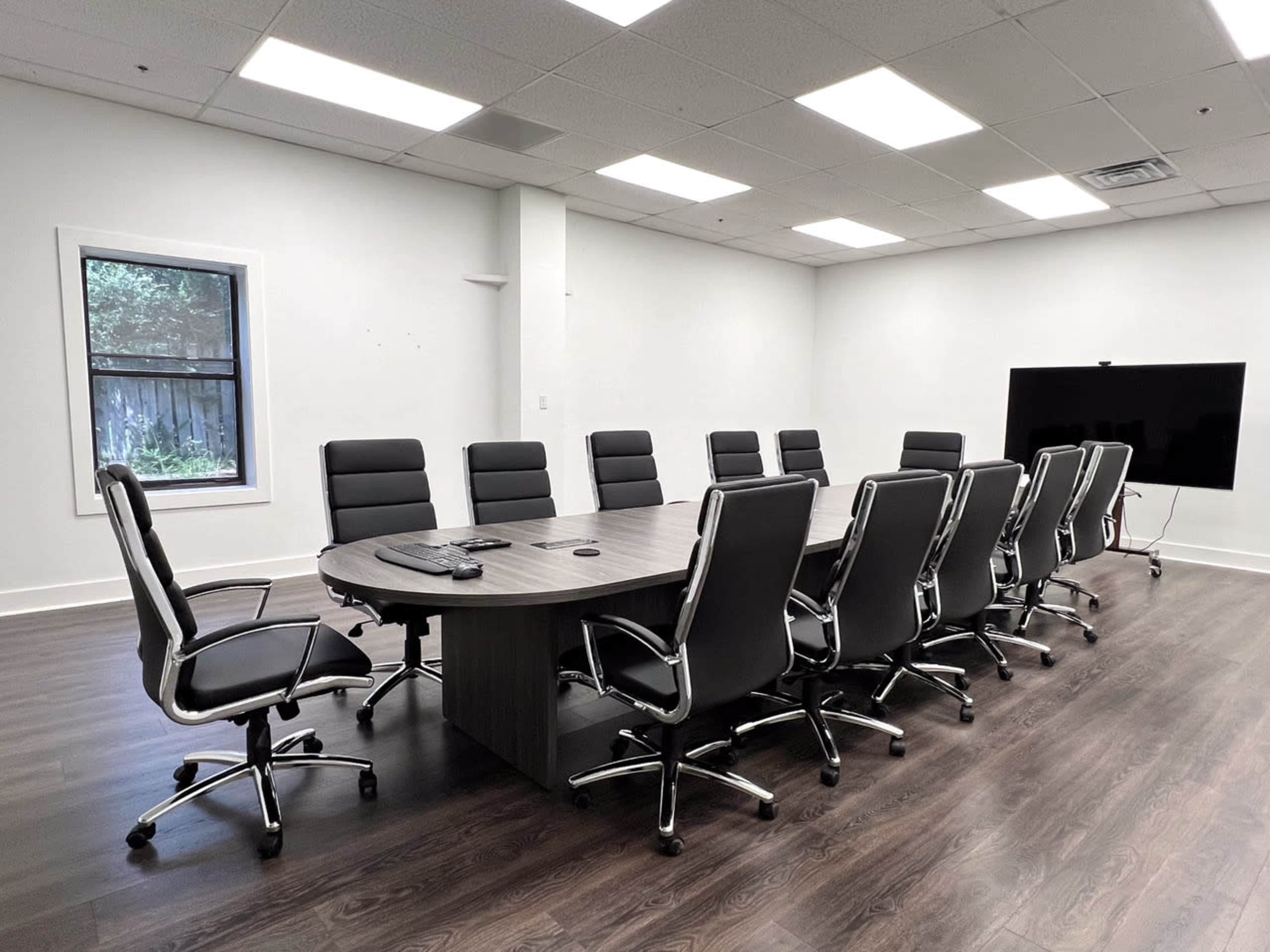 A large conference room features a rectangular table surrounded by 12 black ergonomic chairs, with a flat-screen TV mounted on the wall and a window providing natural light.