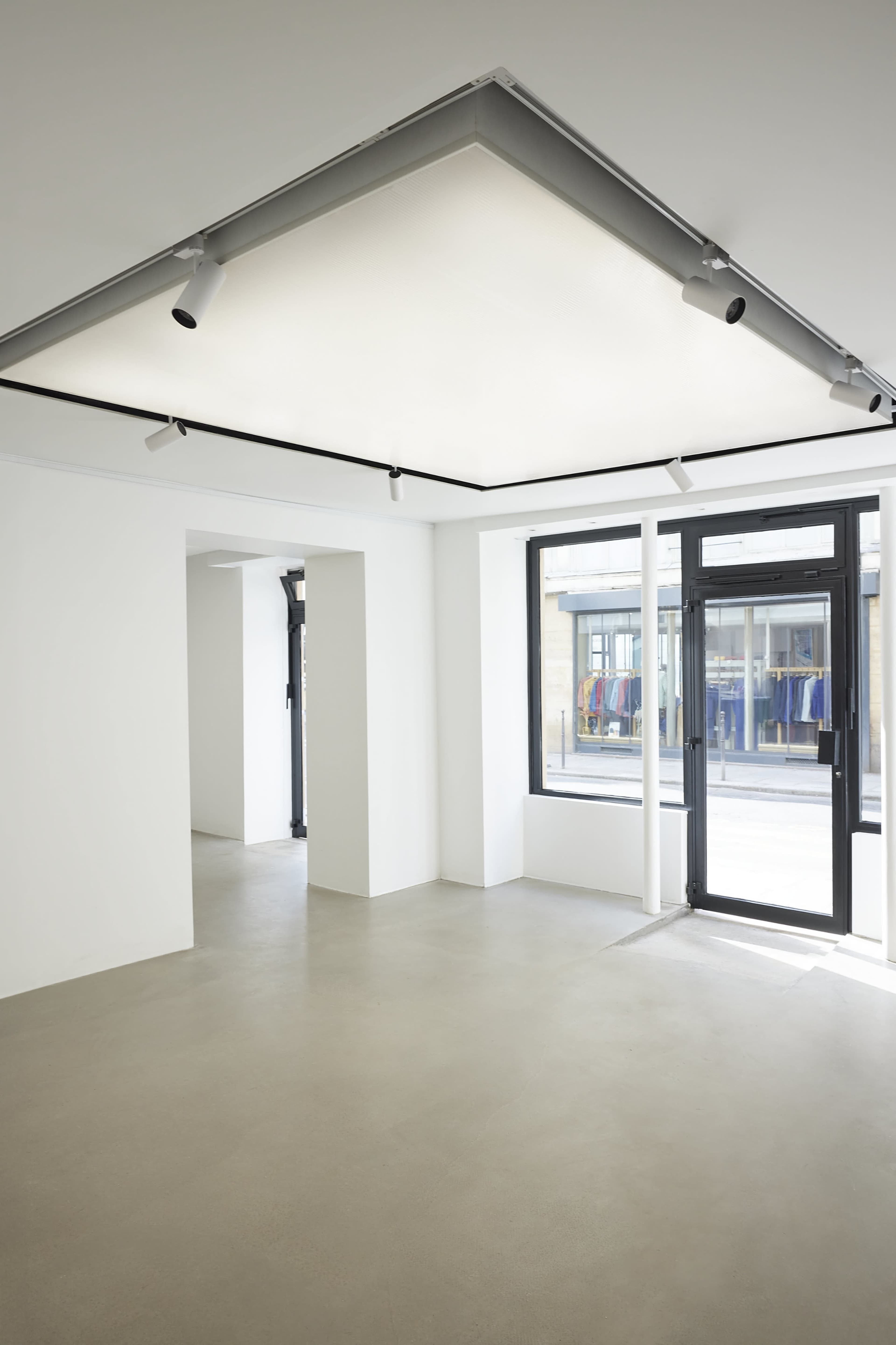 The image shows a spacious, minimalist interior with white walls, a concrete floor, and large windows displaying colorful clothing outside.