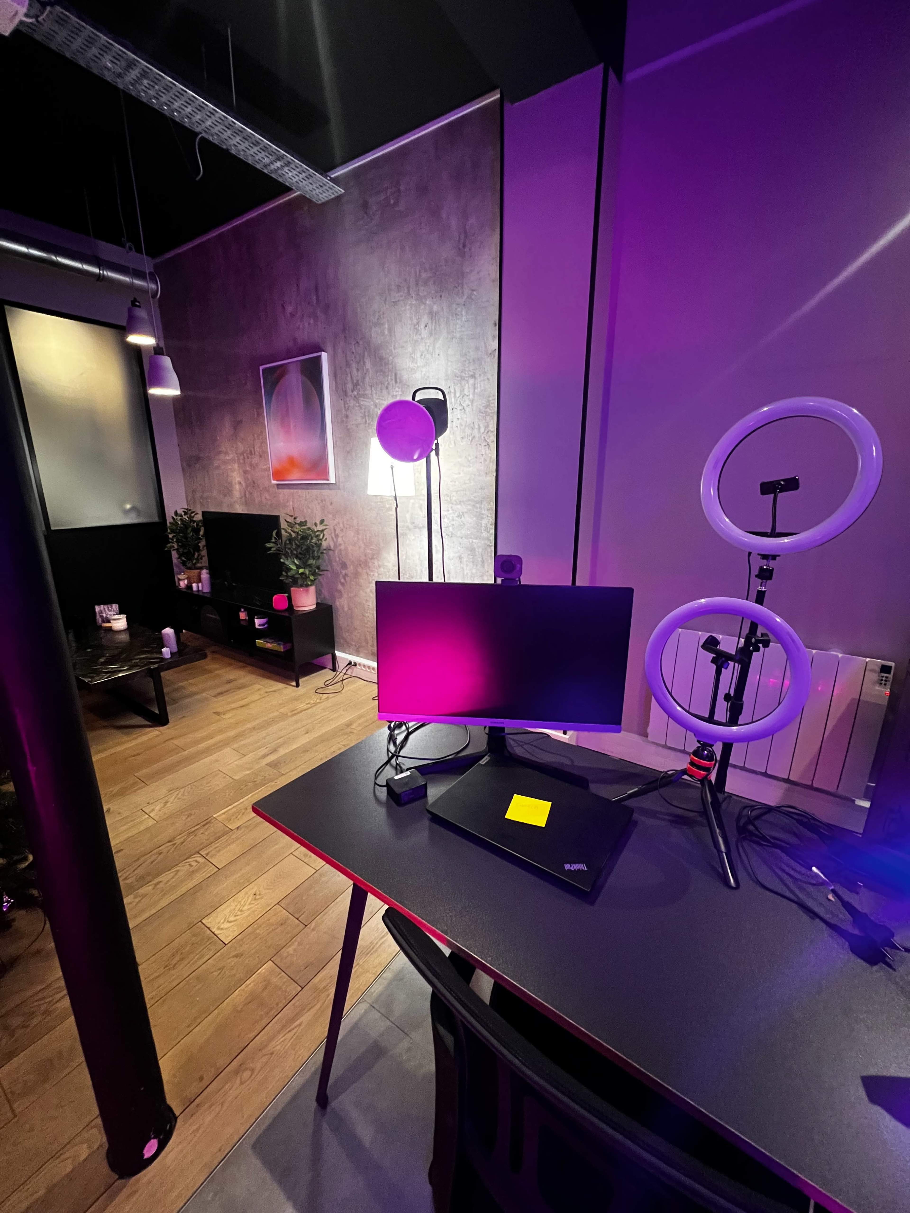 The image shows a modern workspace featuring a monitor on a desk, illuminated by colorful lights and surrounded by potted plants and additional lighting fixtures.