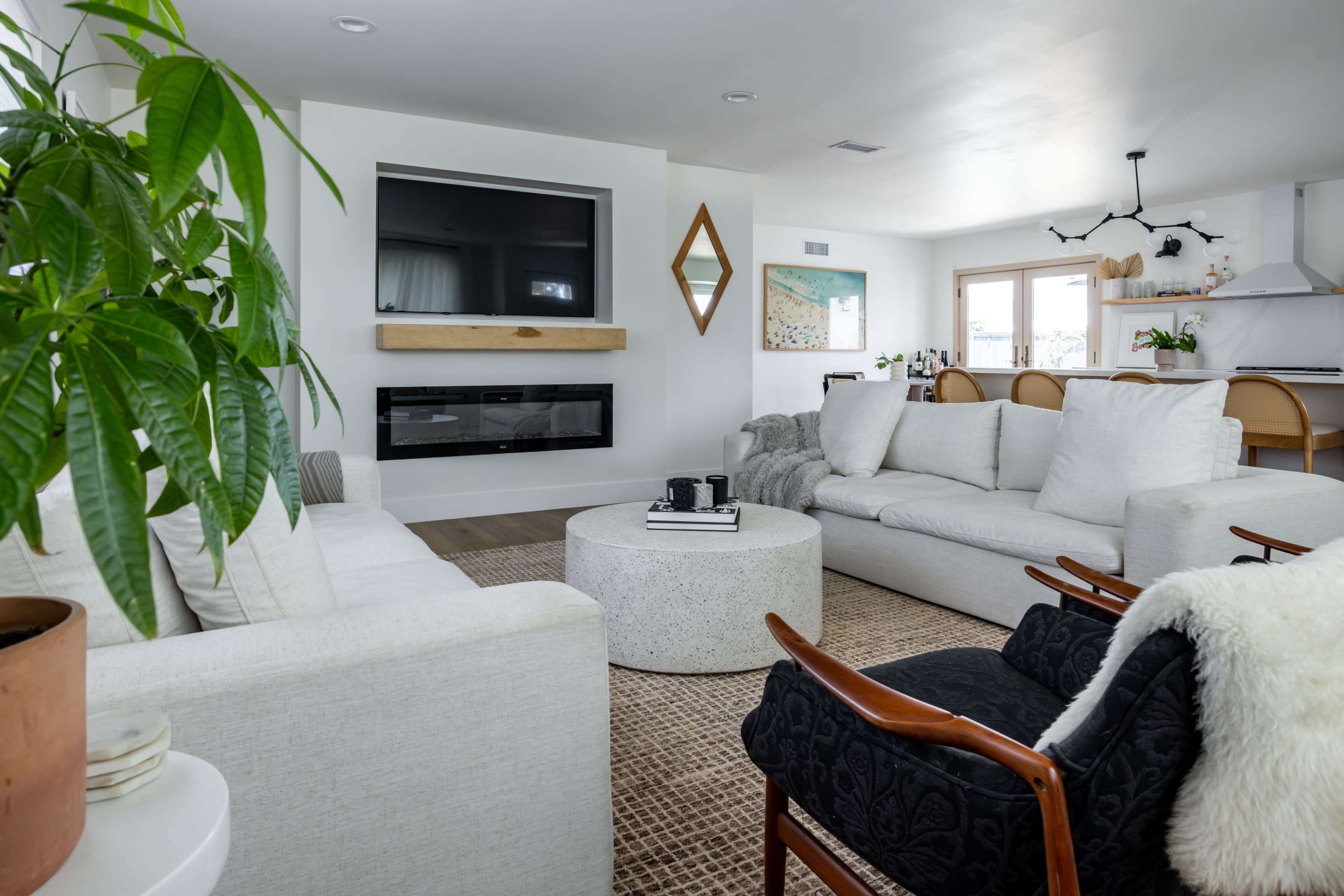 The living room features a modern design with a large television mounted above a fireplace, two white sofas, a round coffee table, and a decorative plant.