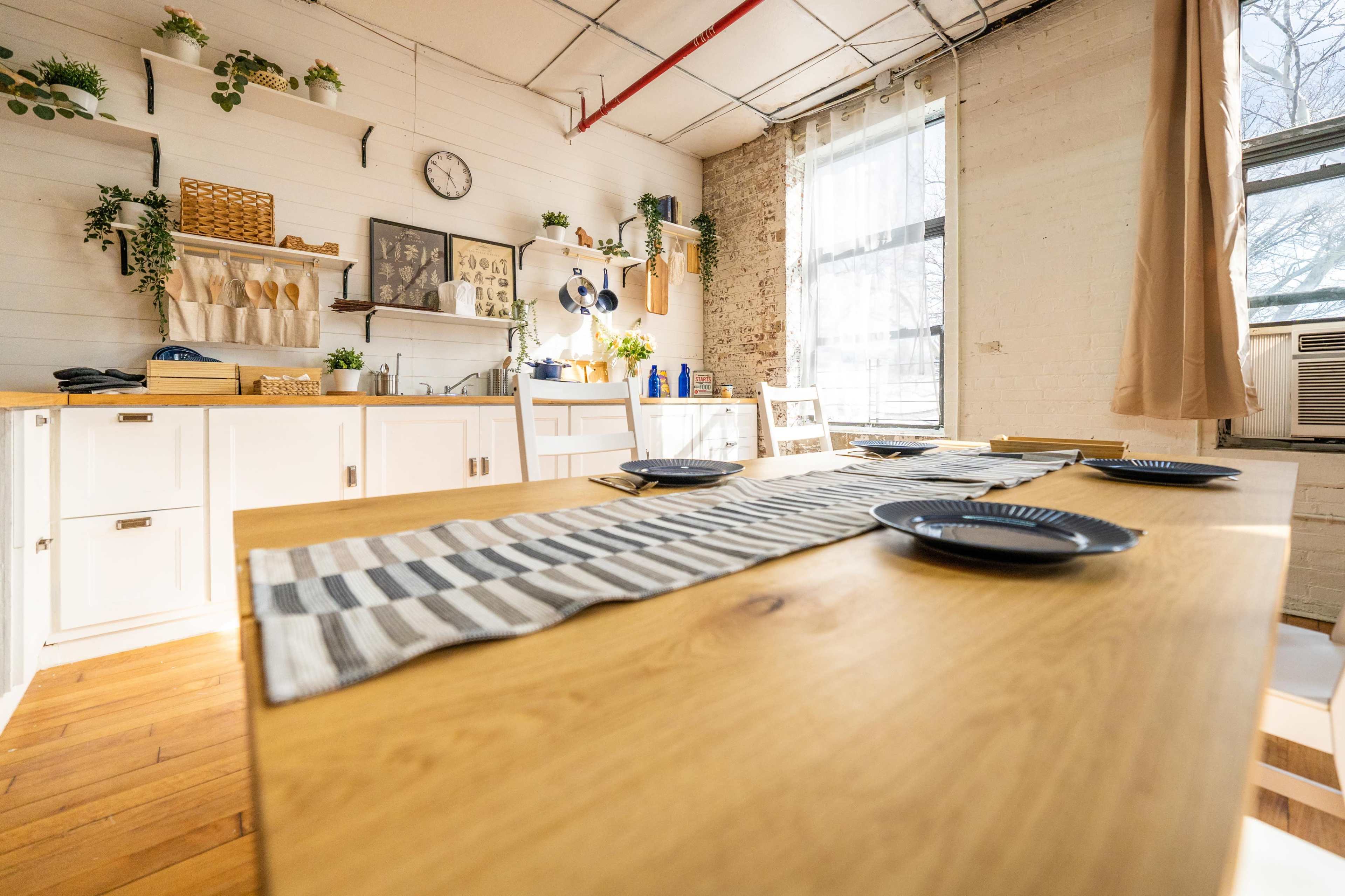 A bright kitchen features a wooden table set for four, surrounded by white cabinets and shelves adorned with utensils and plants.