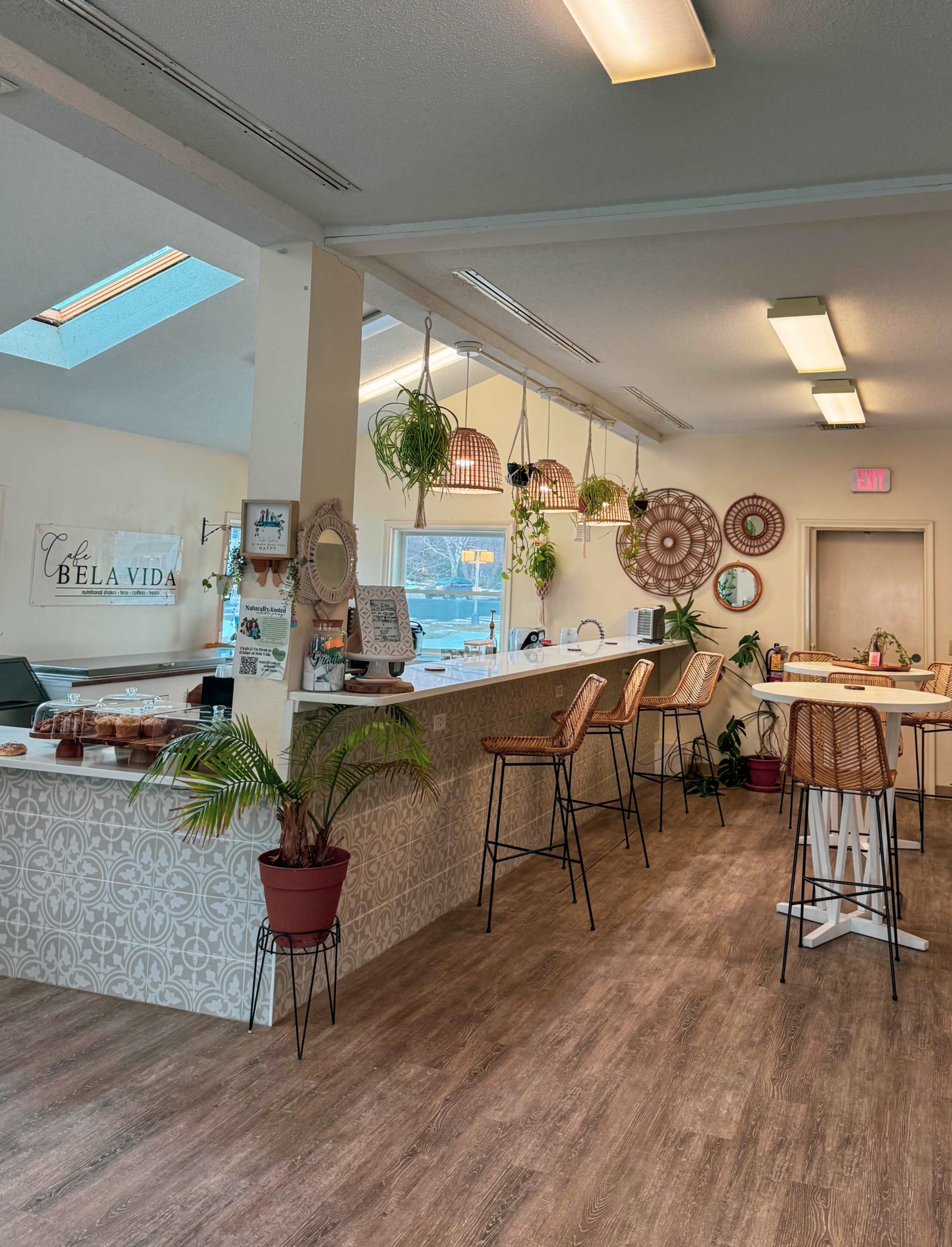 The image shows a cozy café interior with a counter, tall barstools, decorative hanging plants, and a light-colored wooden floor.