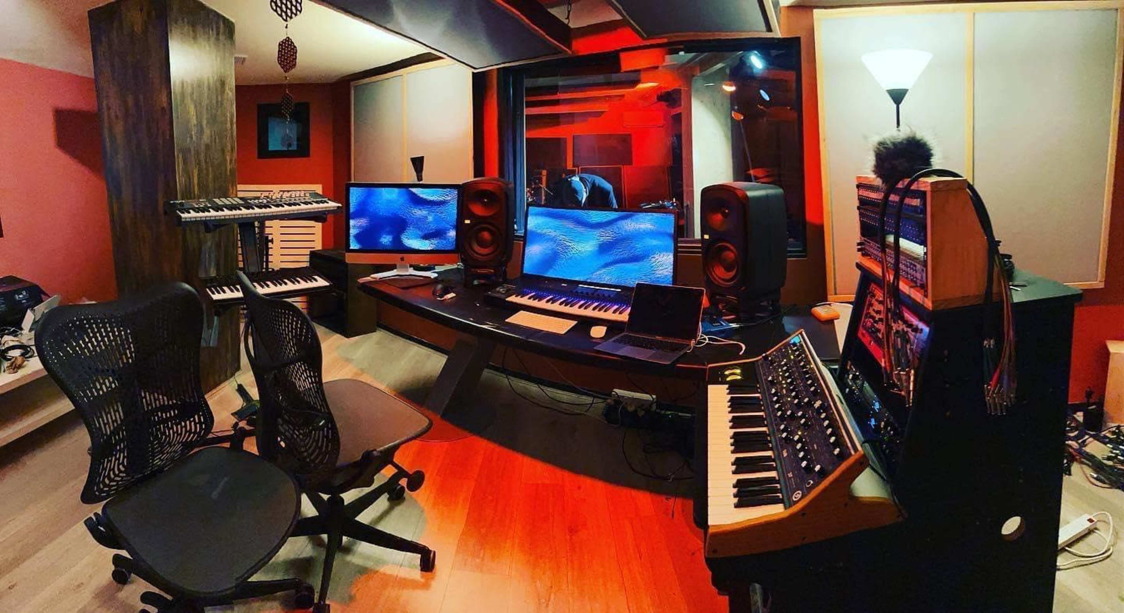 A music studio with electronic instruments, two large monitors, and various audio equipment arranged on a curved desk.