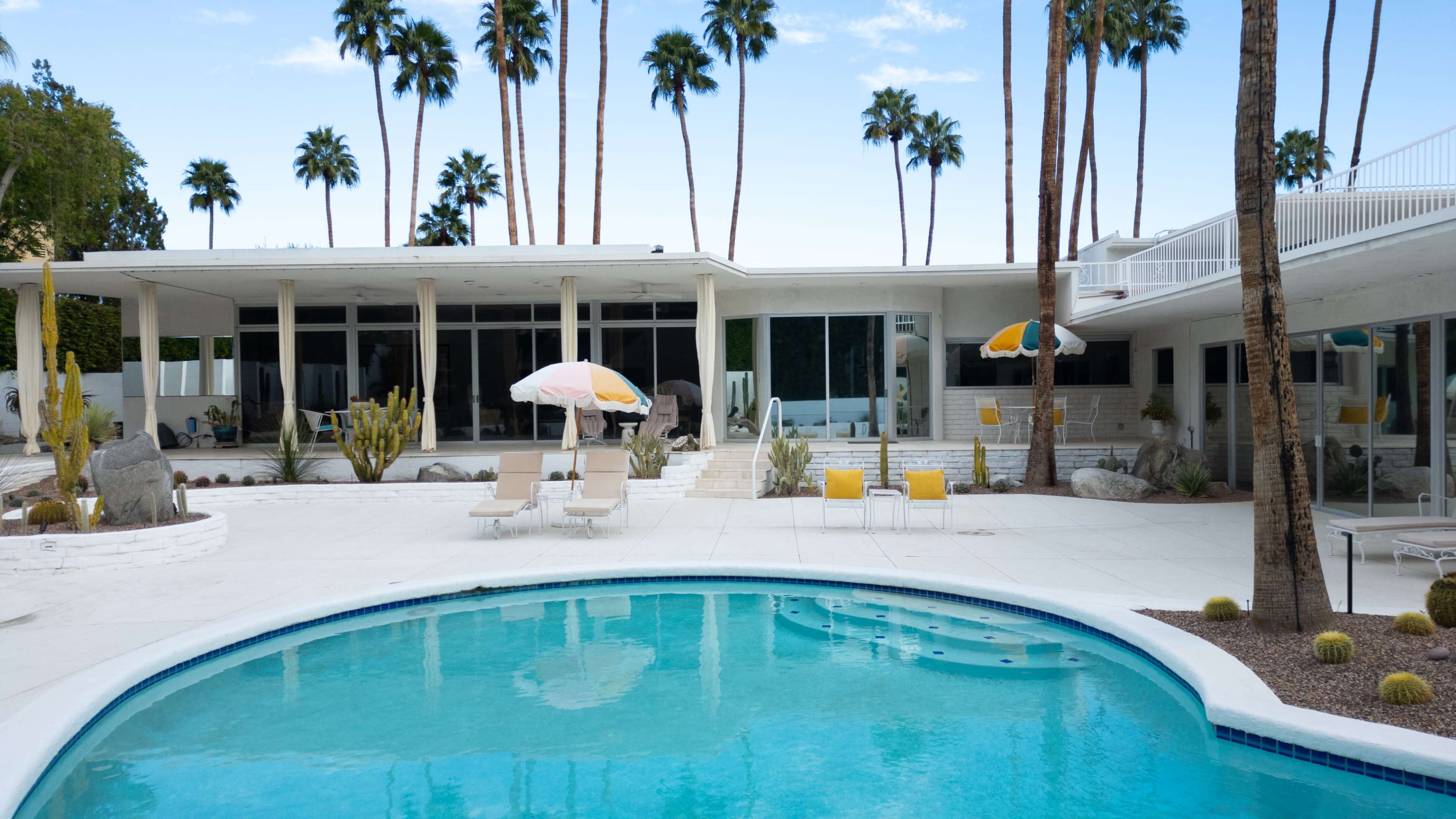 The image shows a modern pool area with a turquoise pool, surrounded by lounge chairs and palm trees against a clear blue sky.